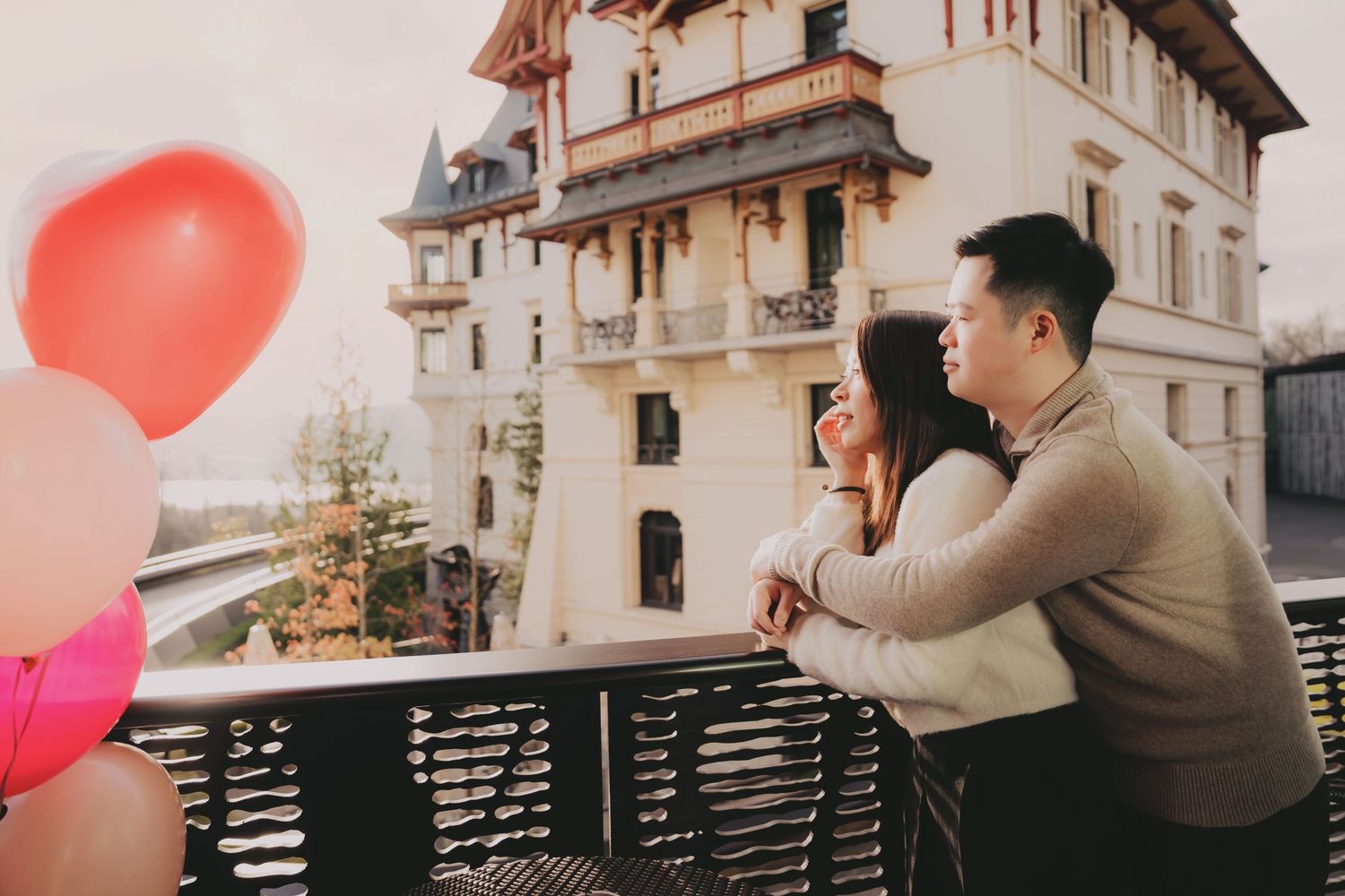 Romantic couple sharing a tender moment on a balcony with pink heart balloons and a European-style building backdrop.