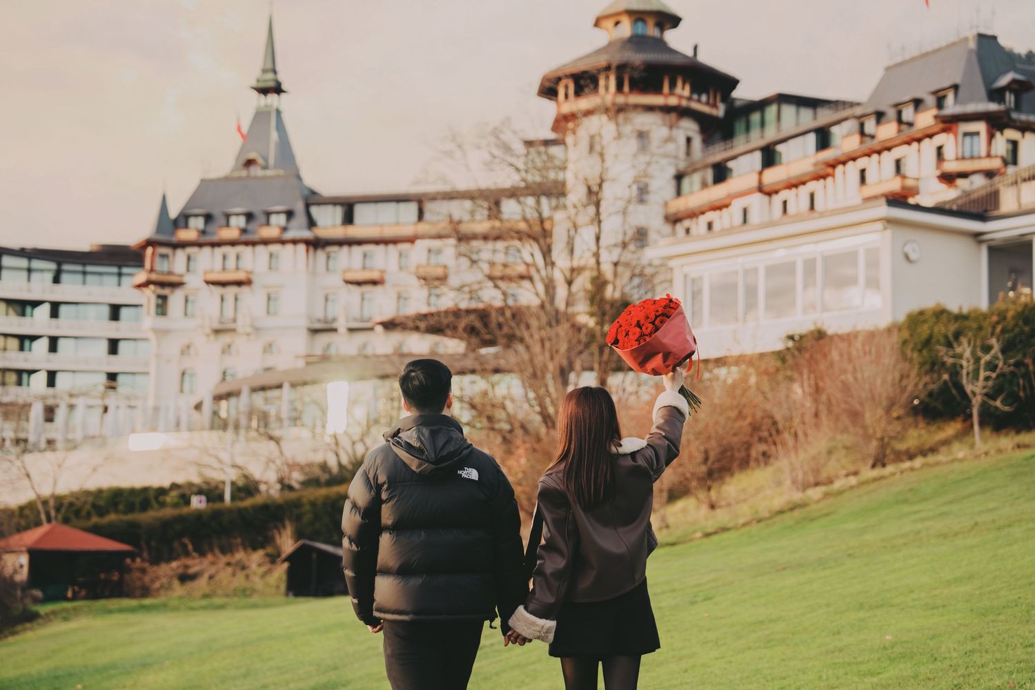Couple holding hands walking toward grand European castle building on lush green lawn.
