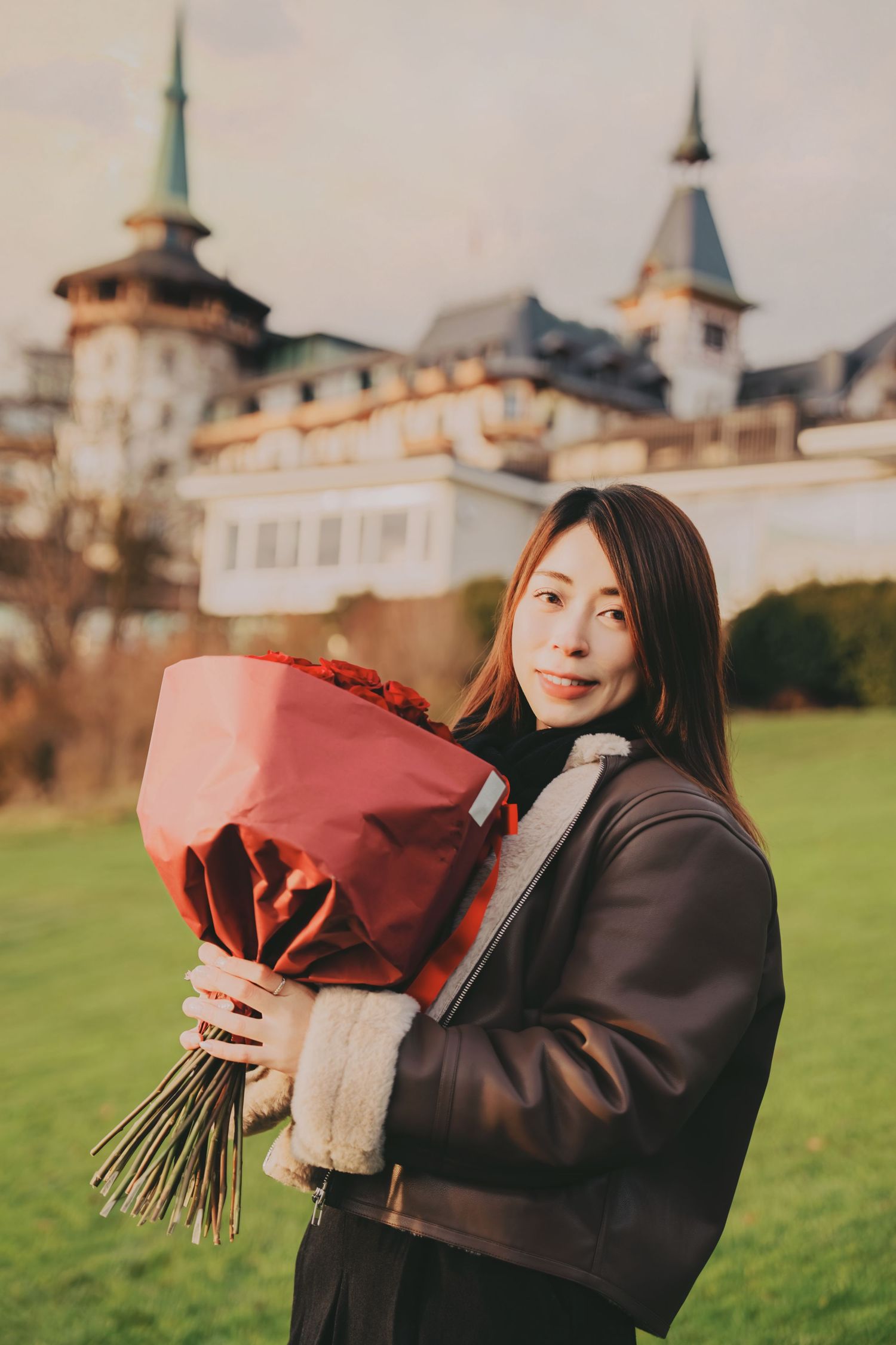 Young Asian woman holding large red rose bouquet standing before historic castle towers.