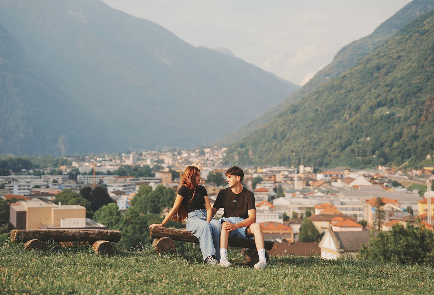 Young couple sitting together on a bench overlooking a scenic valley town surrounded by mountains.