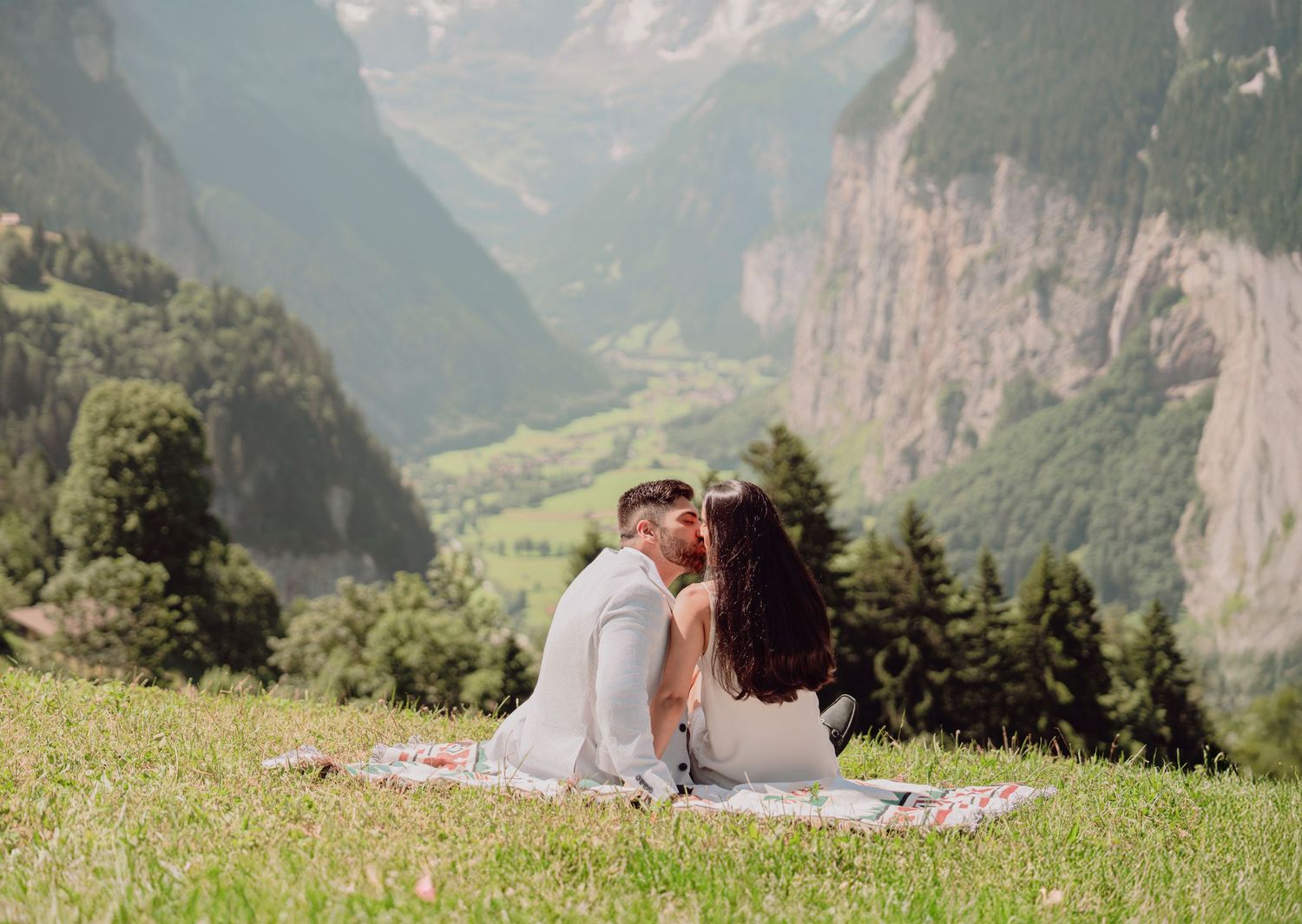 Couple sharing a kiss while seated on a blanket on a grassy slope overlooking a scenic Swiss mountain valley.