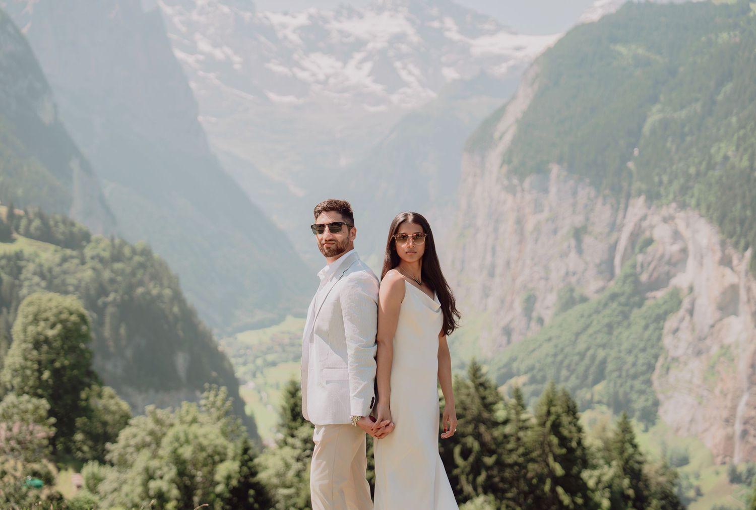 Elegant couple holding hands and posing together with a stunning Swiss alpine valley landscape behind them.