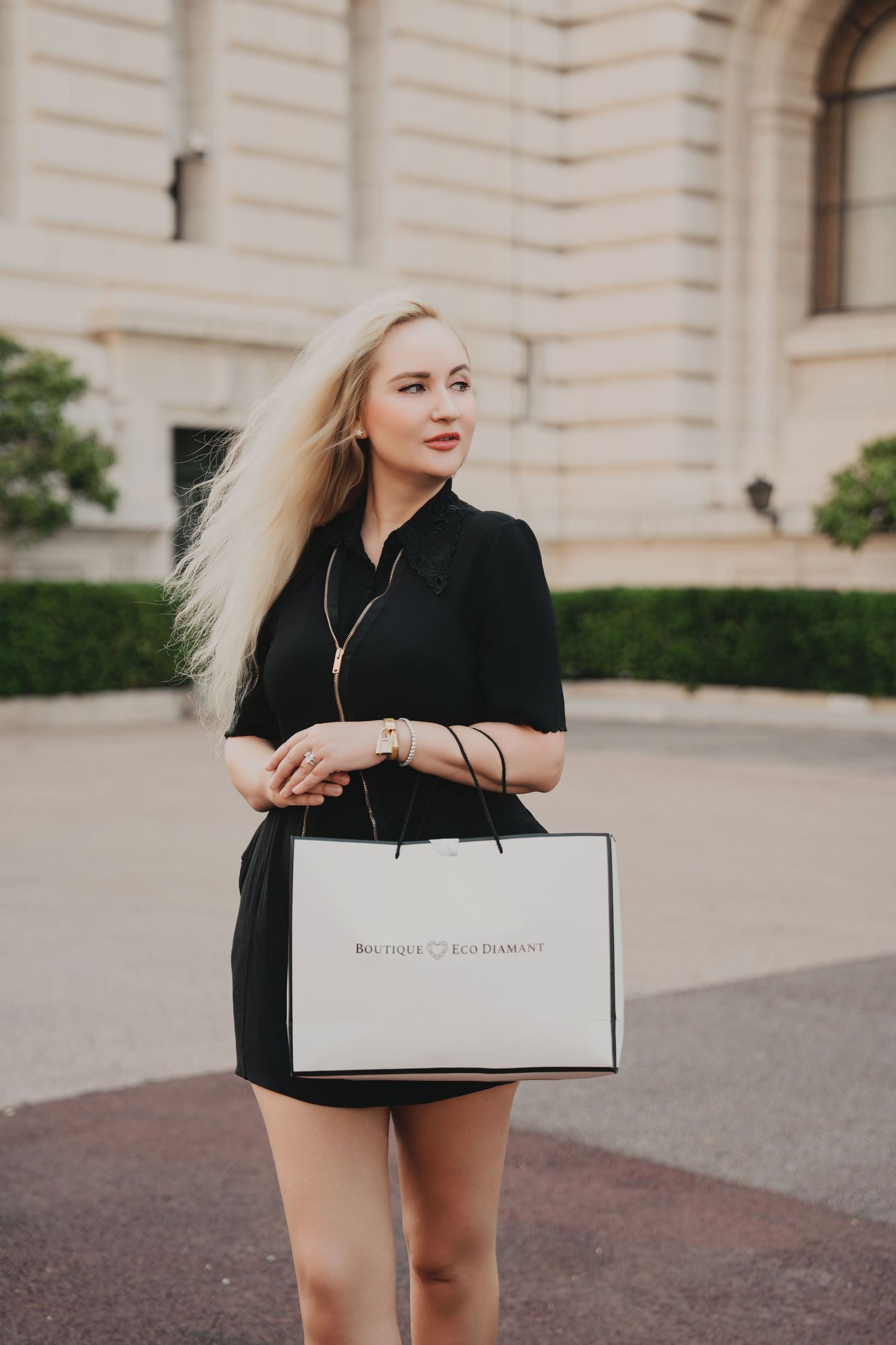 Smiling blonde woman in black dress holding a boutique shopping bag on a European street.