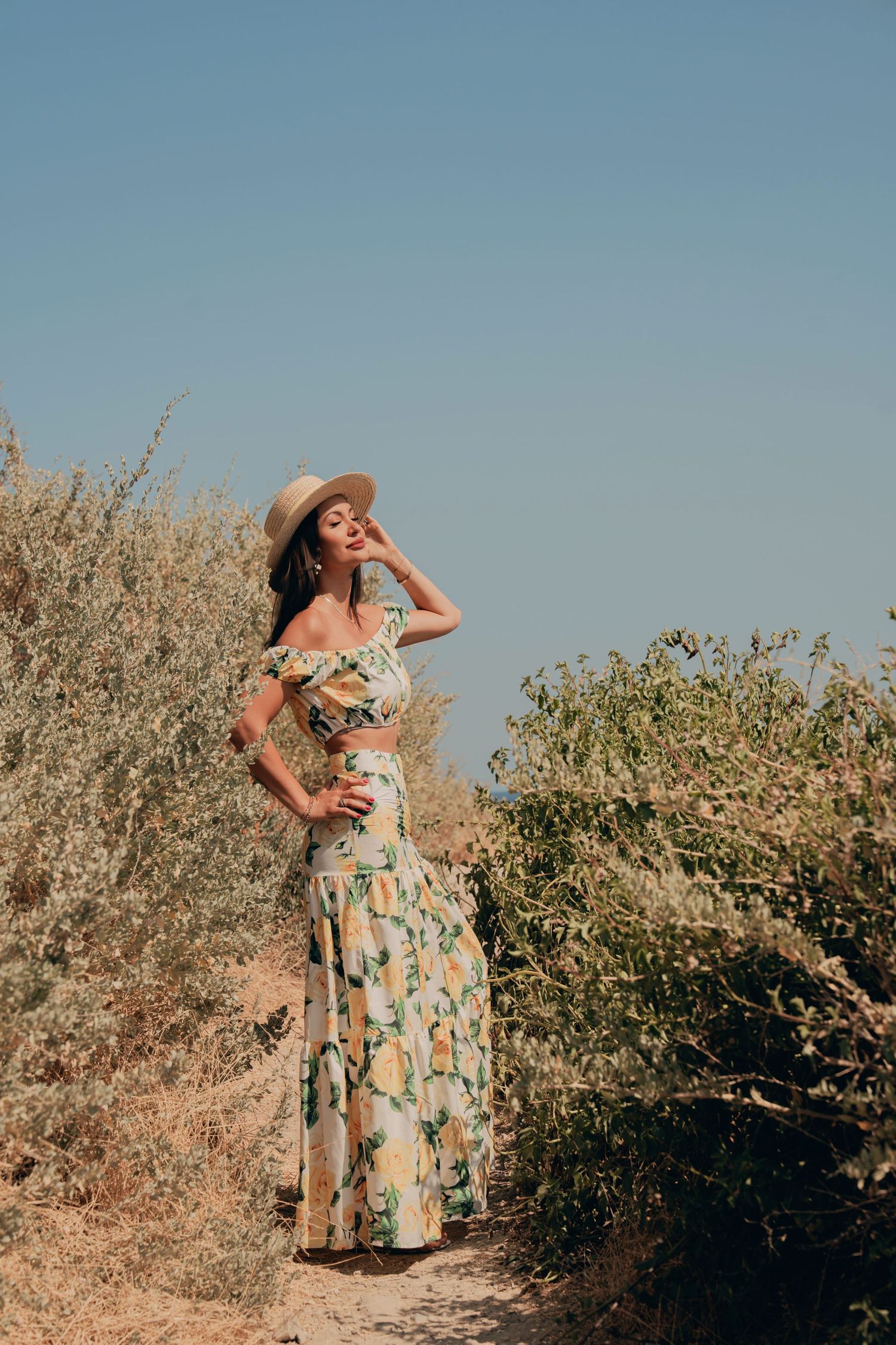 Elegant woman in floral maxi dress and straw hat posing among sun-dried Mediterranean shrubs.