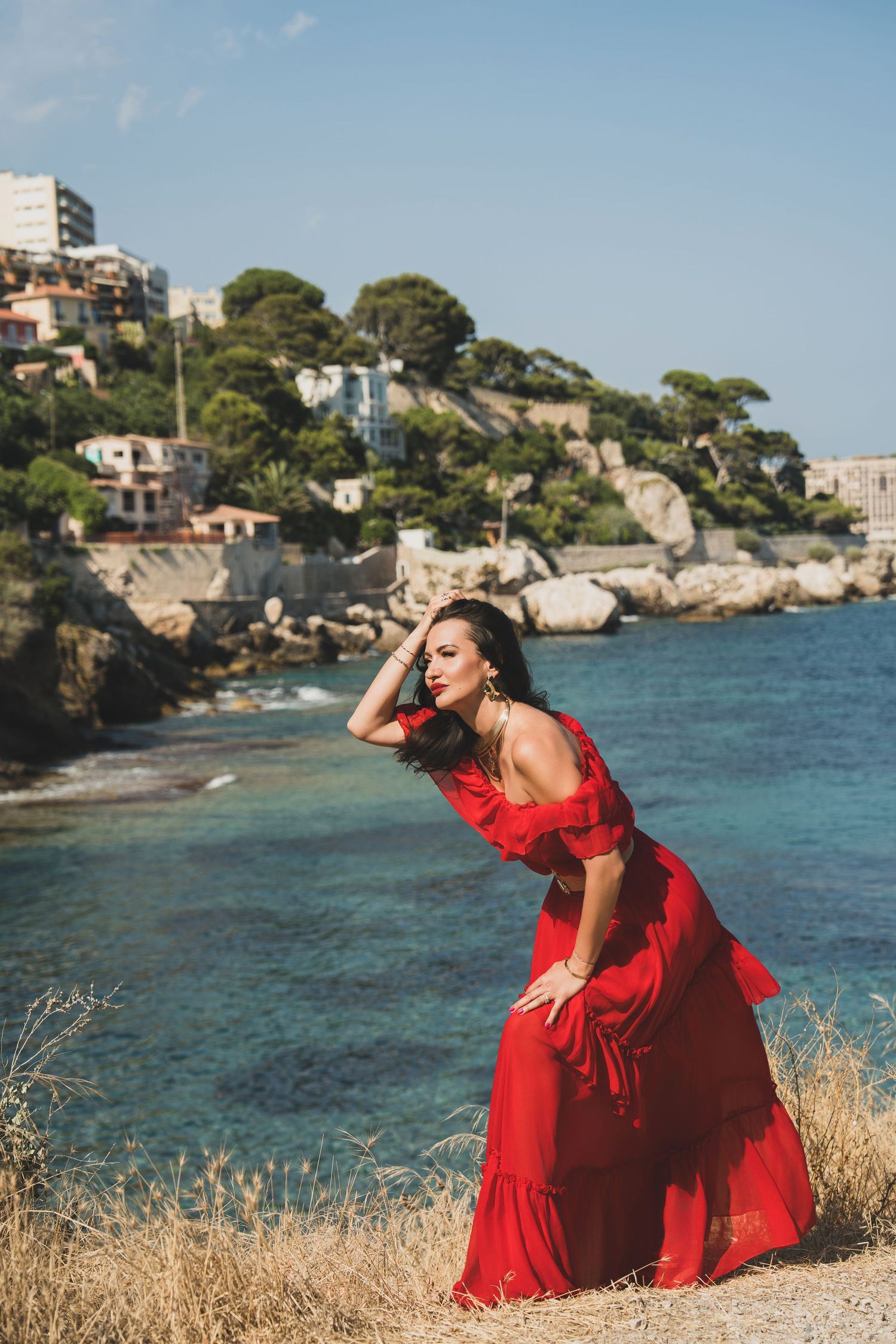 Brunette woman in red maxi dress posing on rocky seaside cliff with scenic coastal town behind.