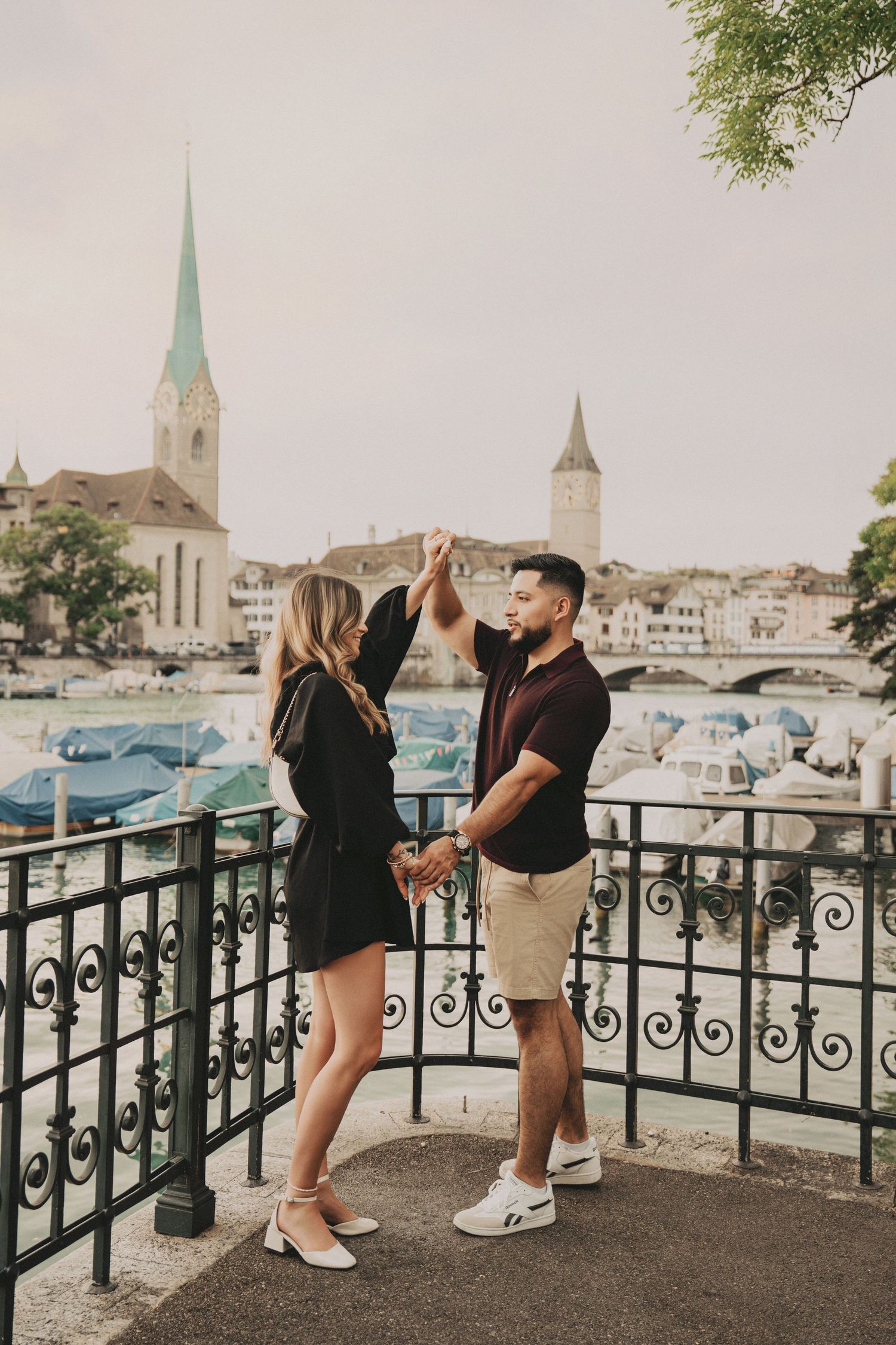 Romantic couple spinning together on an ornate balcony with Zurich cityscape and river behind them.