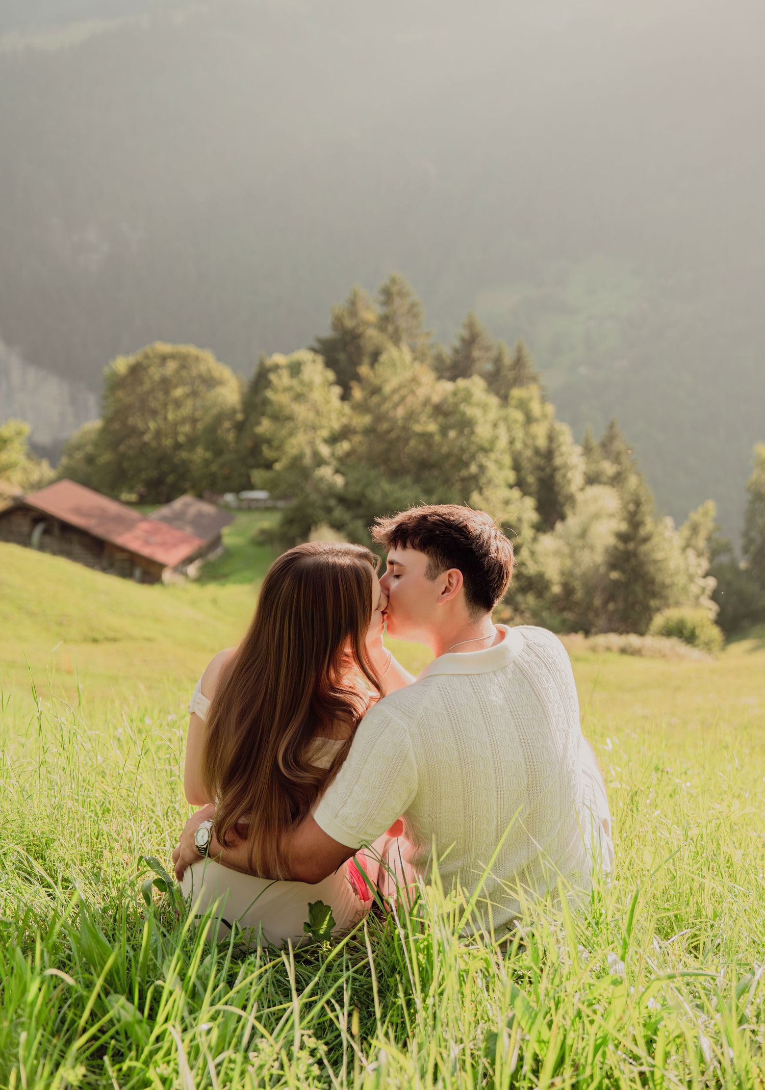 Couple sitting close together in green alpine meadow sharing a kiss with mountain backdrop.