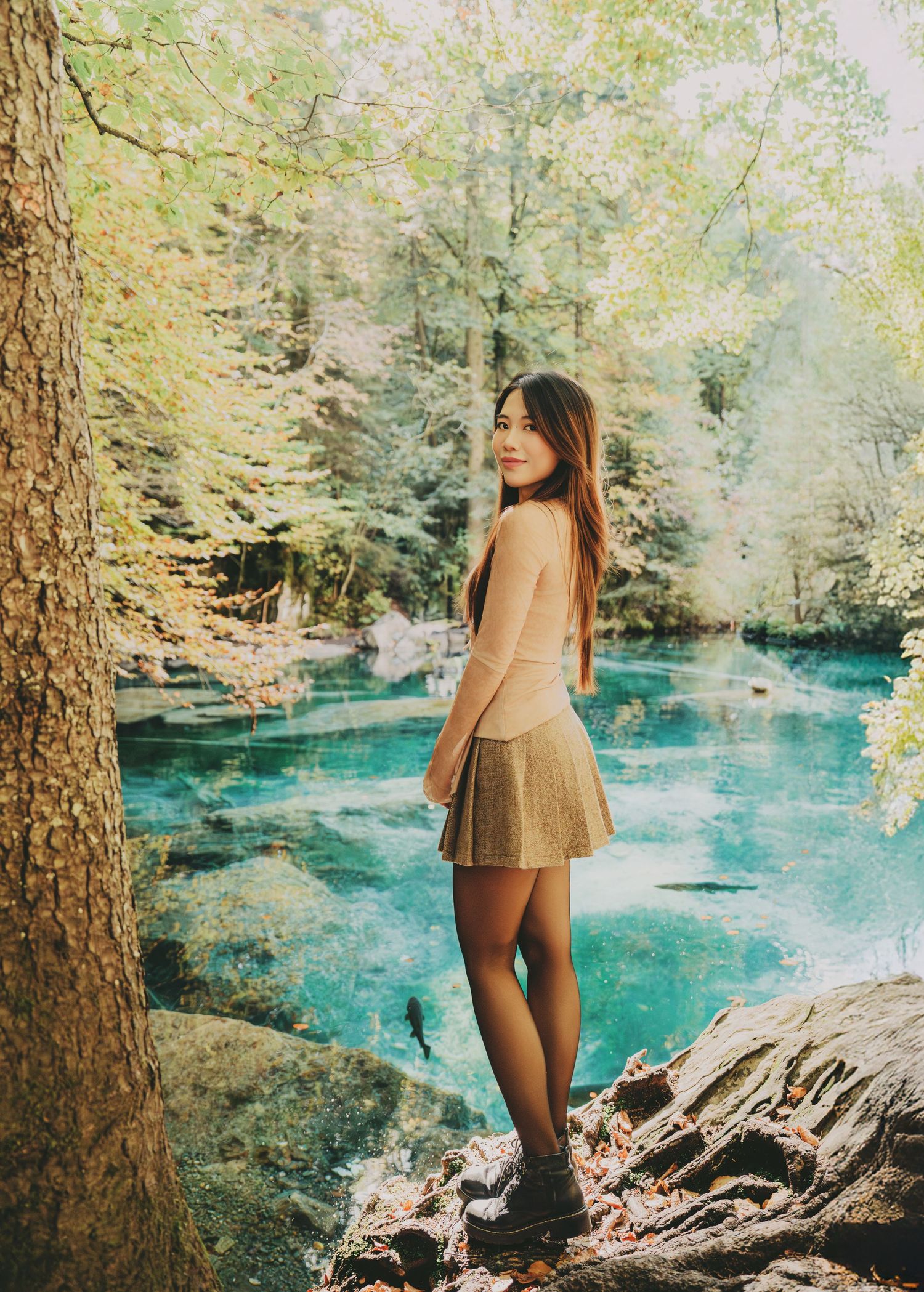Smiling woman in beige dress poses by a crystal clear turquoise river surrounded by lush green trees.