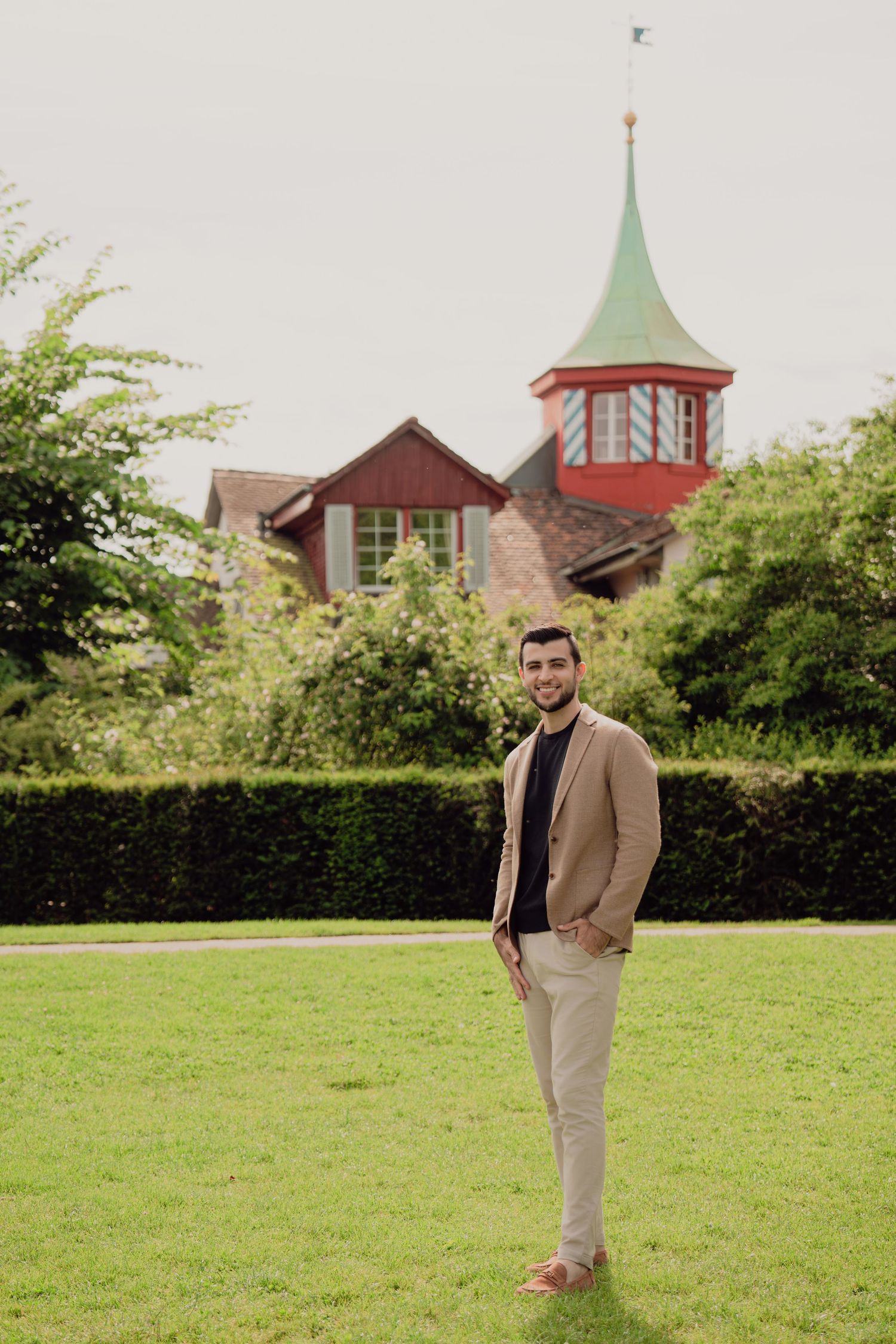 Smiling man in beige blazer and khakis standing on green lawn with red-spired building behind him.