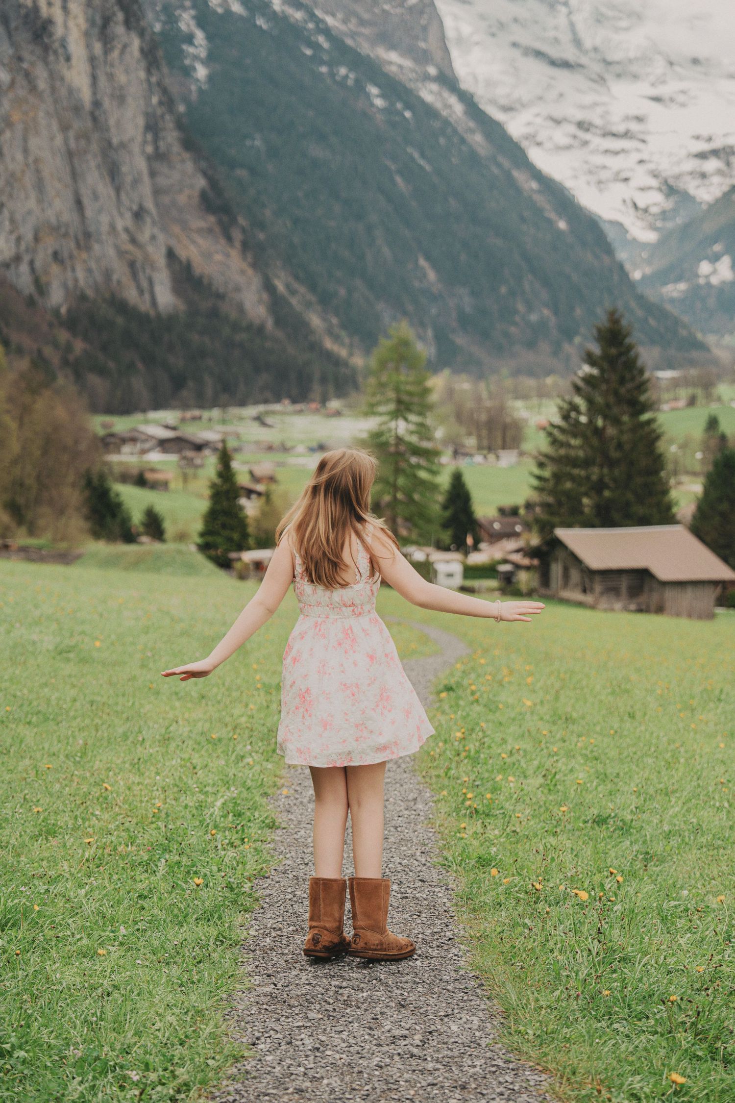Young woman in floral dress and brown boots standing with arms outstretched on a path overlooking a lush Alpine valley.