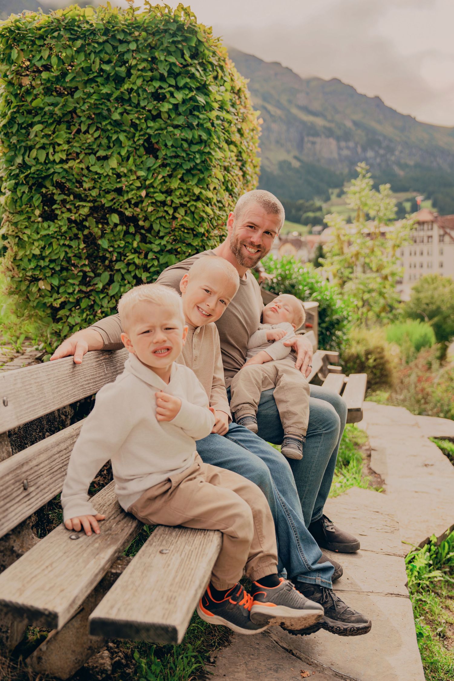 Father sitting on a bench with three young sons in a scenic Swiss mountain setting.