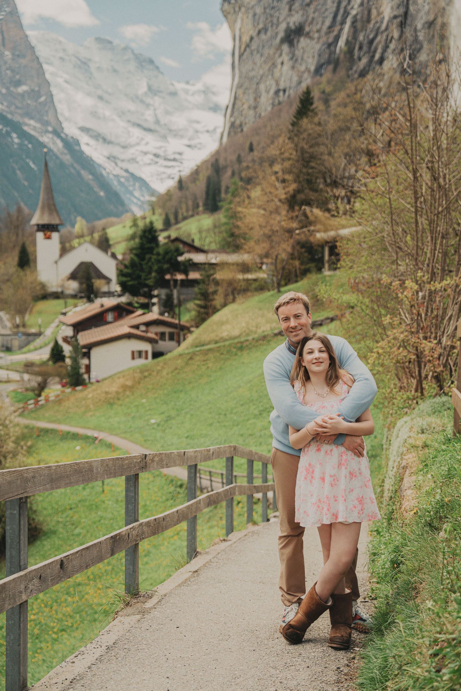 A couple poses affectionately on a mountain path with a picturesque Swiss village and snowy peaks behind them.