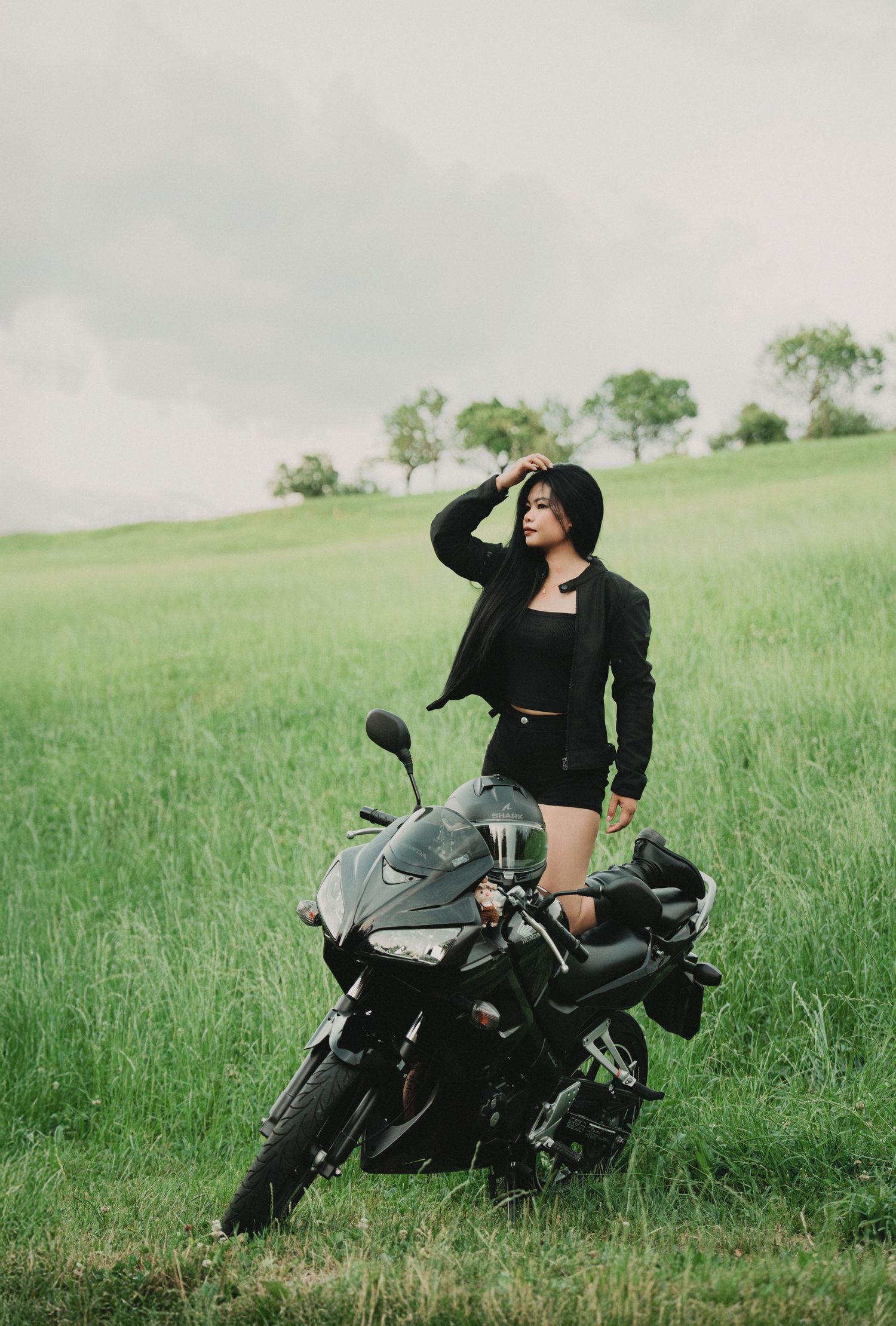 Stylish woman in black shorts and jacket standing beside a black motorcycle in a green field.