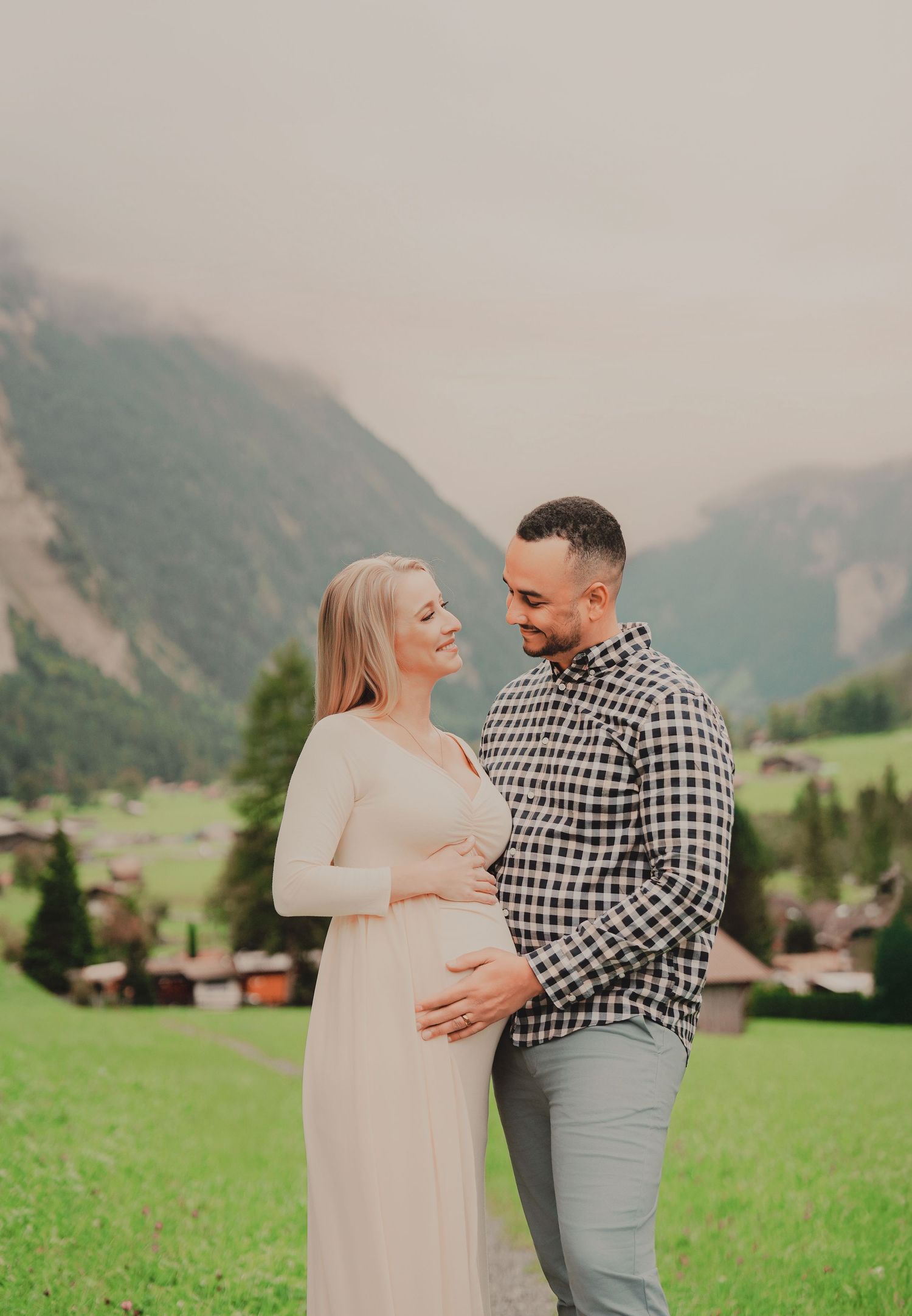 Smiling expecting couple holding baby bump in green alpine meadow with misty Swiss mountain backdrop.