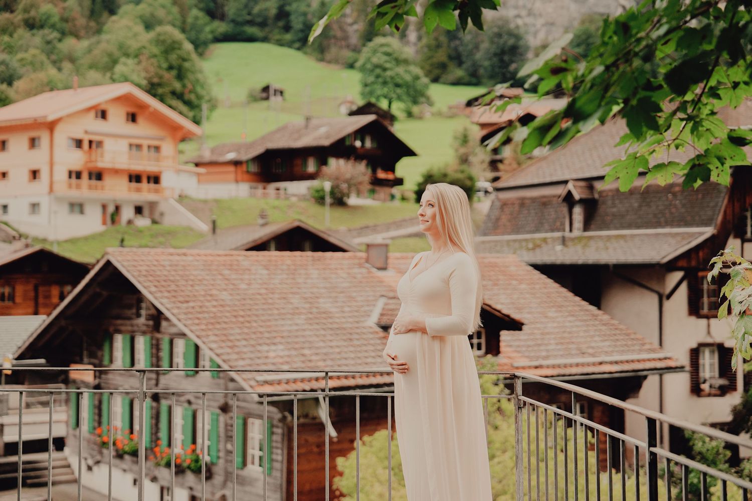 Pregnant woman in white dress on balcony overlooking Swiss alpine village with traditional wooden chalets.