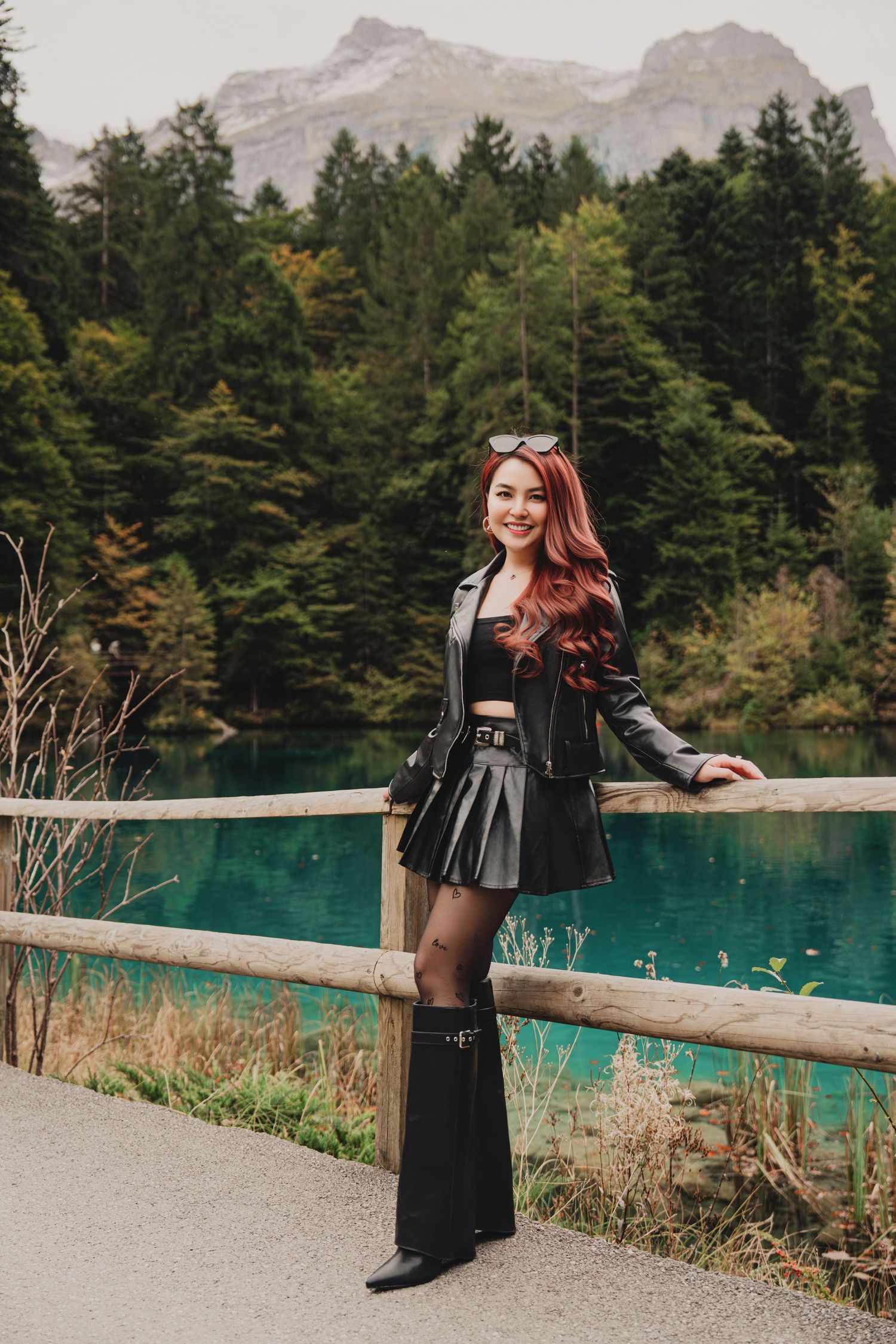 Red-haired woman in black leather outfit posing by wooden fence with turquoise lake and mountains.