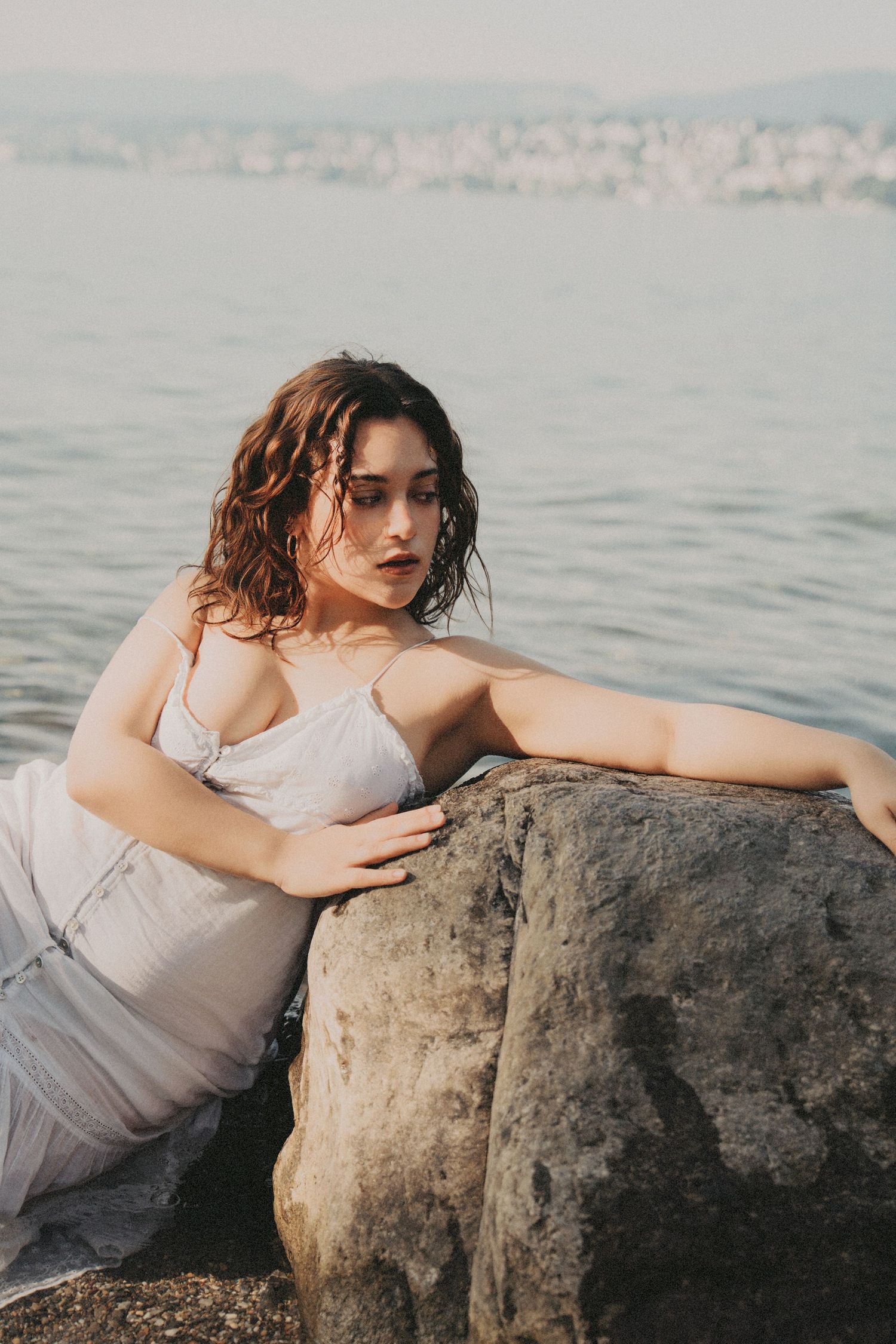 Woman in white dress leaning on seaside rock with city skyline in background.