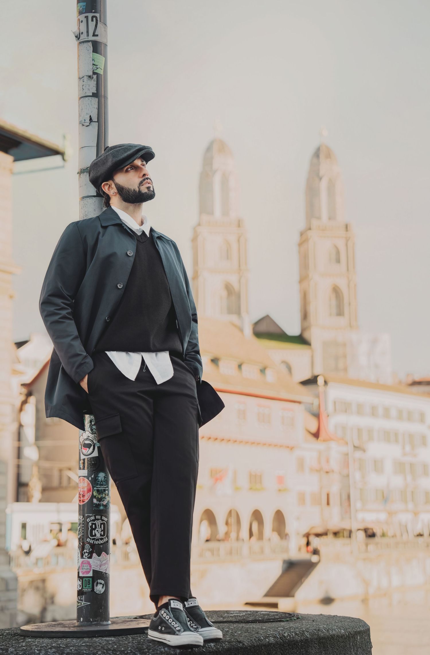 Man in black coat, flat cap, and sneakers standing on a stone block with Zurich Grossmunster in background.