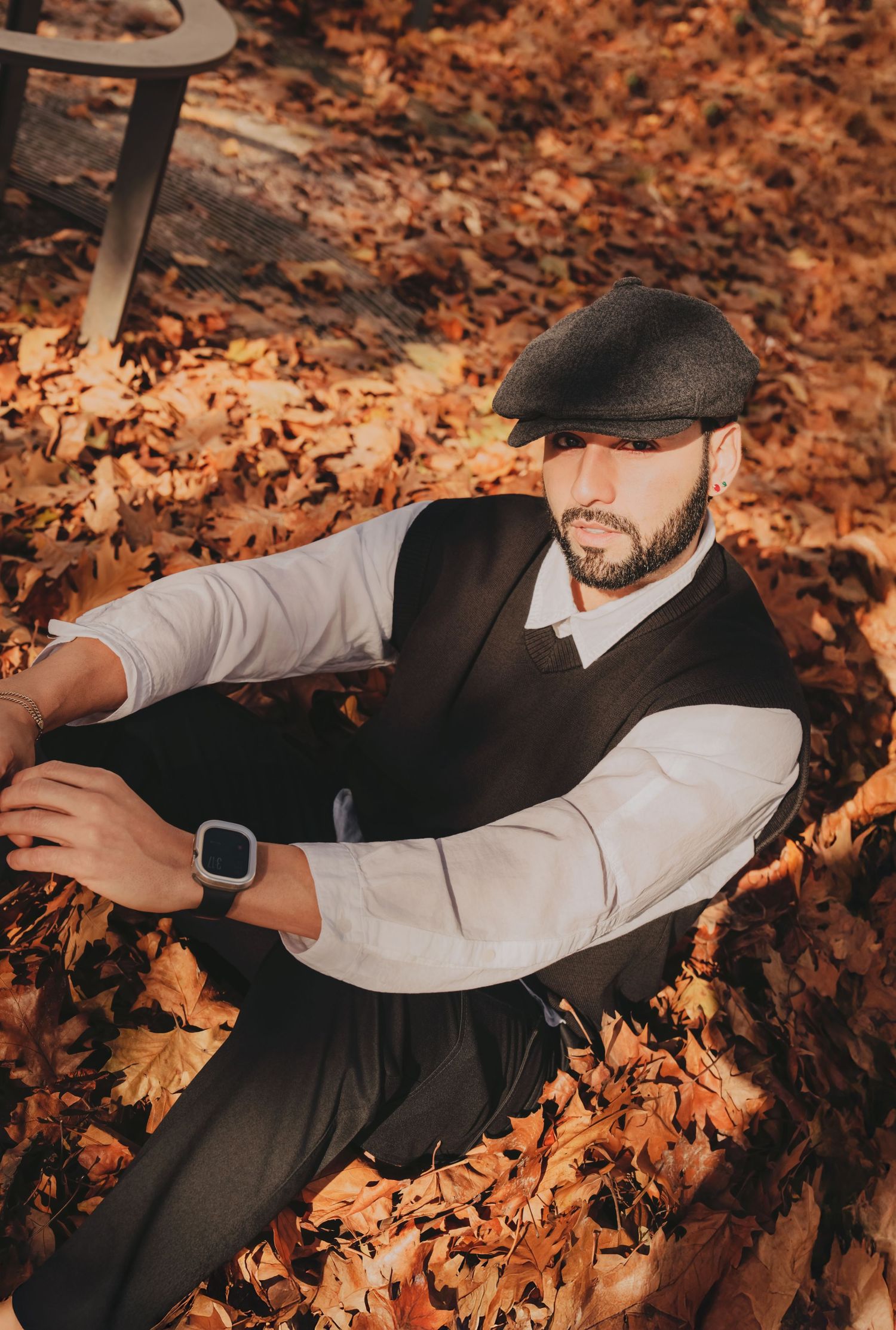 Man in white shirt and vest sitting in pile of golden autumn leaves wearing flat cap.