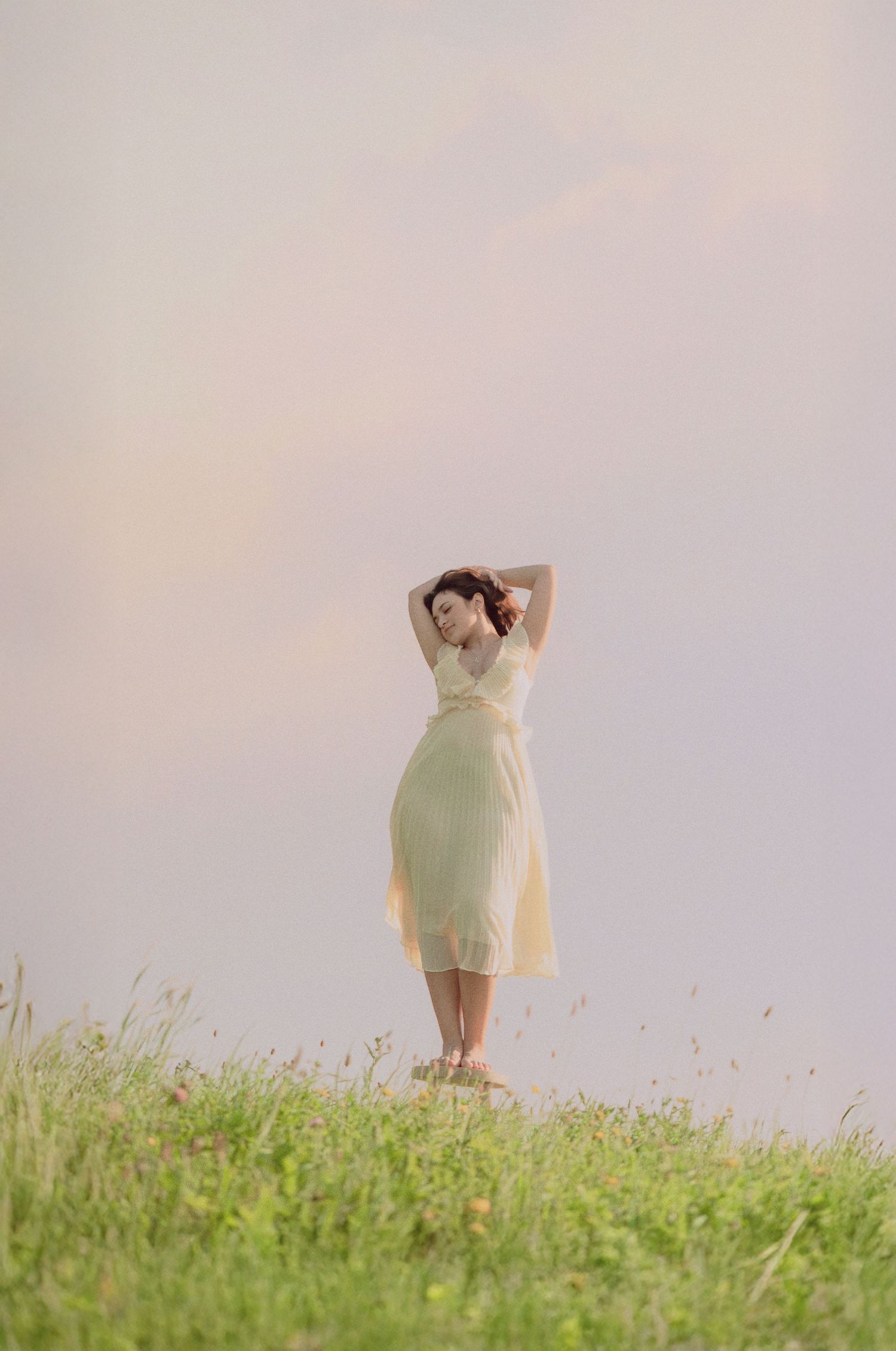 Woman in flowing yellow dress standing in wildflower meadow with hands behind head under soft sky.