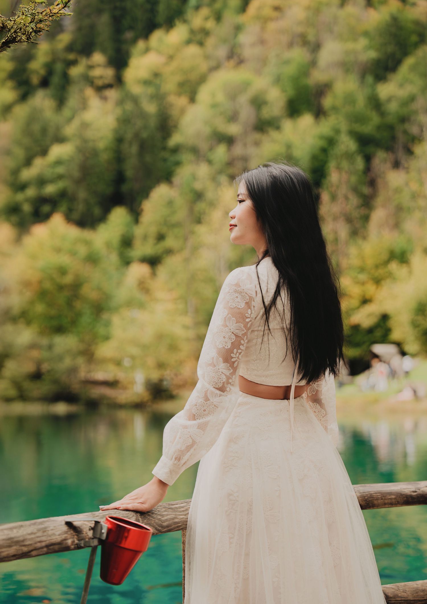 Woman in white lace crop top and skirt holds red cup by scenic turquoise lake.