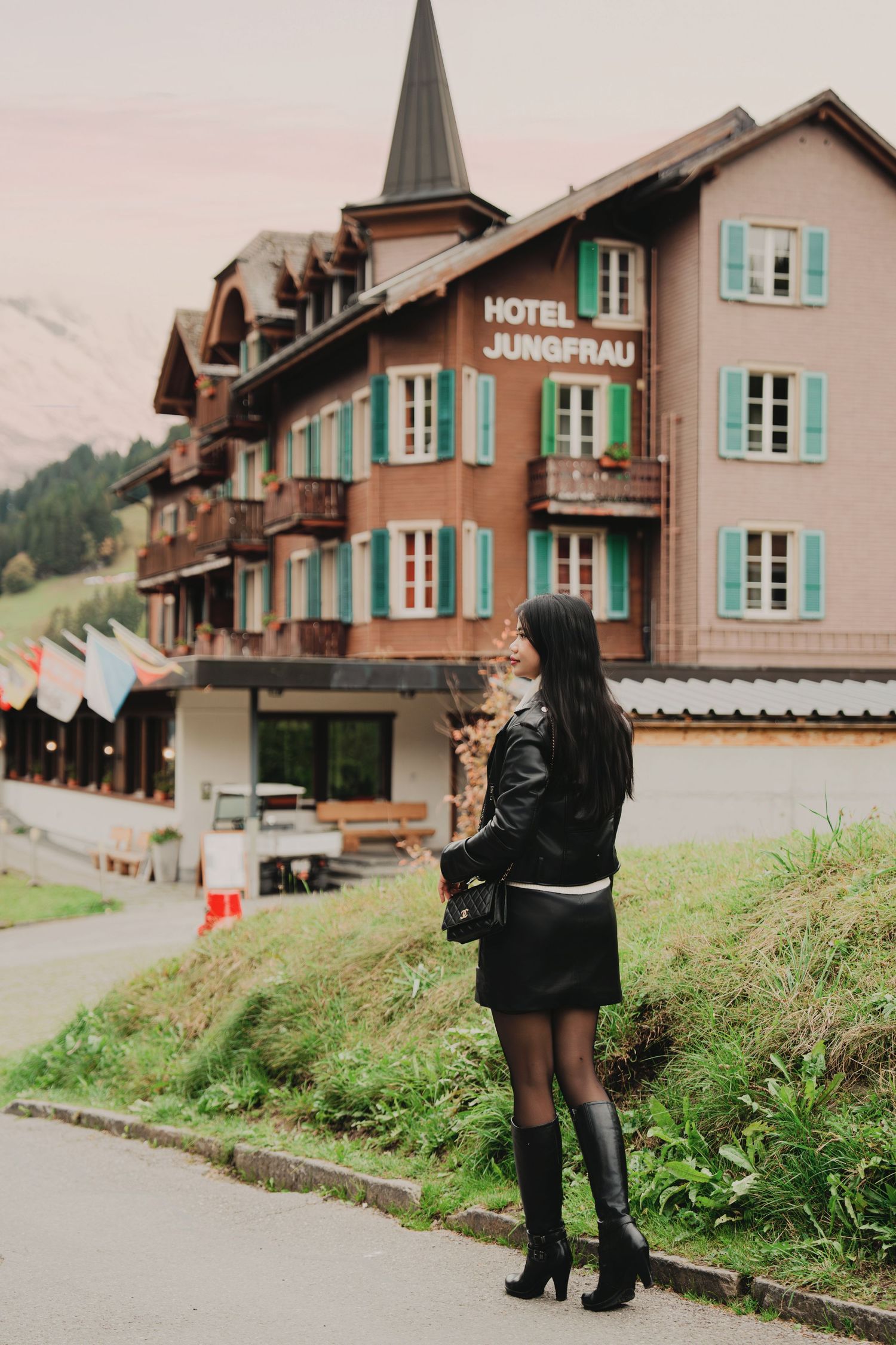 Woman in black leather jacket poses in front of Hotel Jungfrau building in Swiss mountain village.