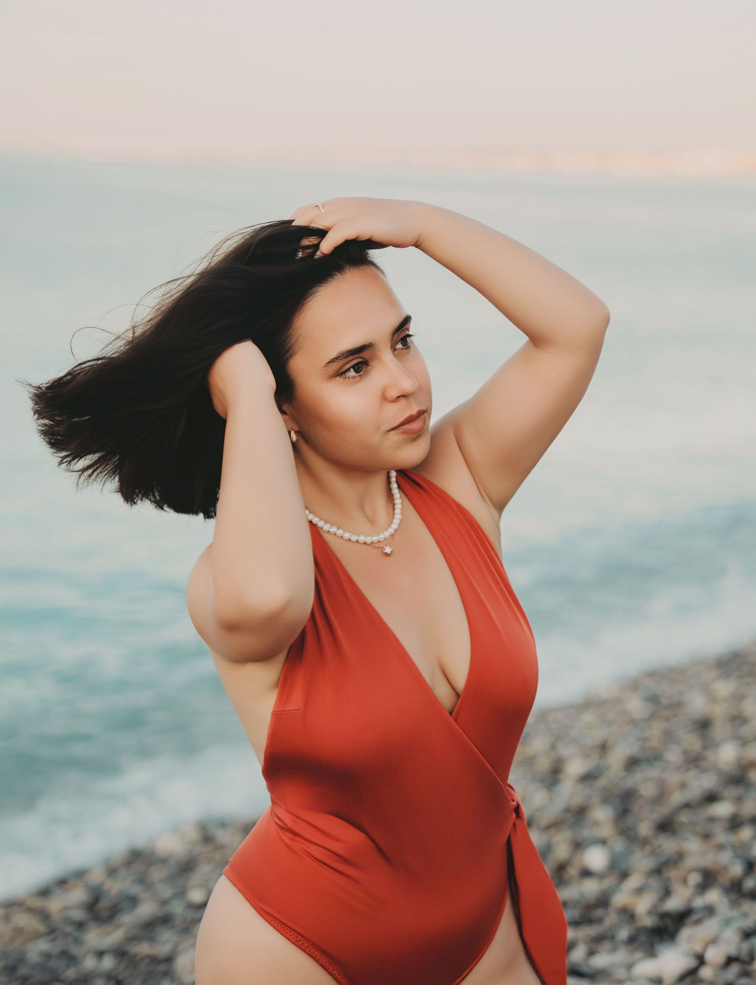 Young brunette woman in red halter swimsuit and pearl necklace posing on pebble beach at sunset.