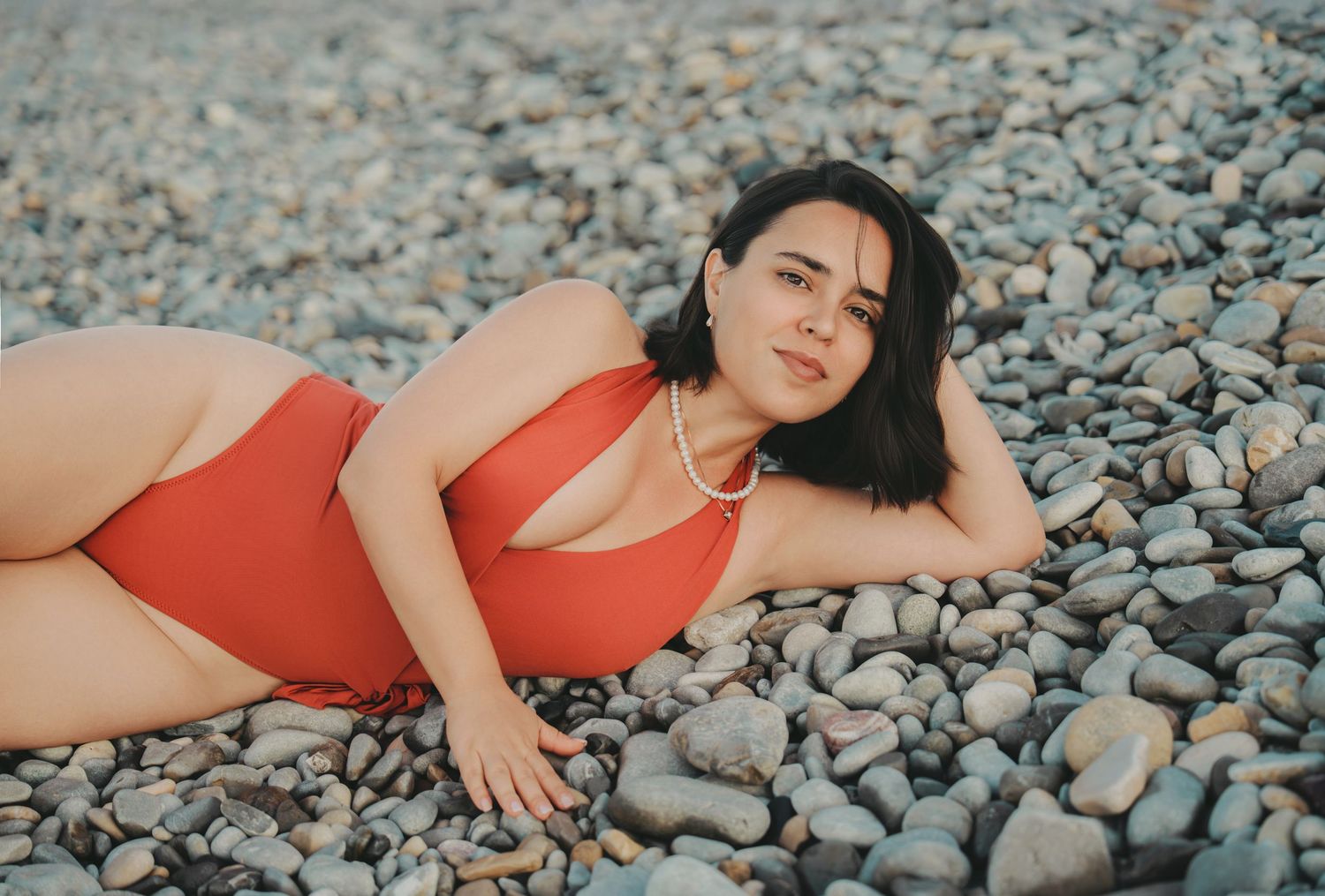 A woman in a red swimsuit reclines on a rocky pebble beach by the ocean.