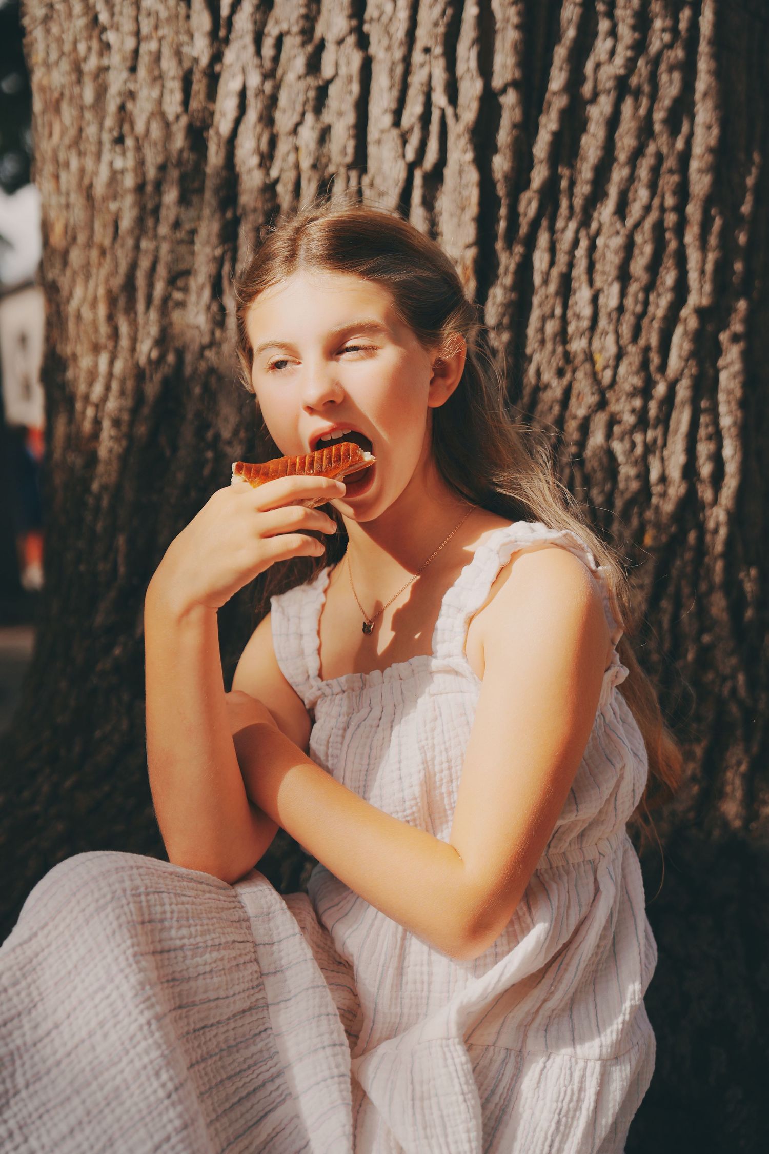 A young girl in a white dress sits against a tree trunk, eating a chocolate cupcake outdoors.