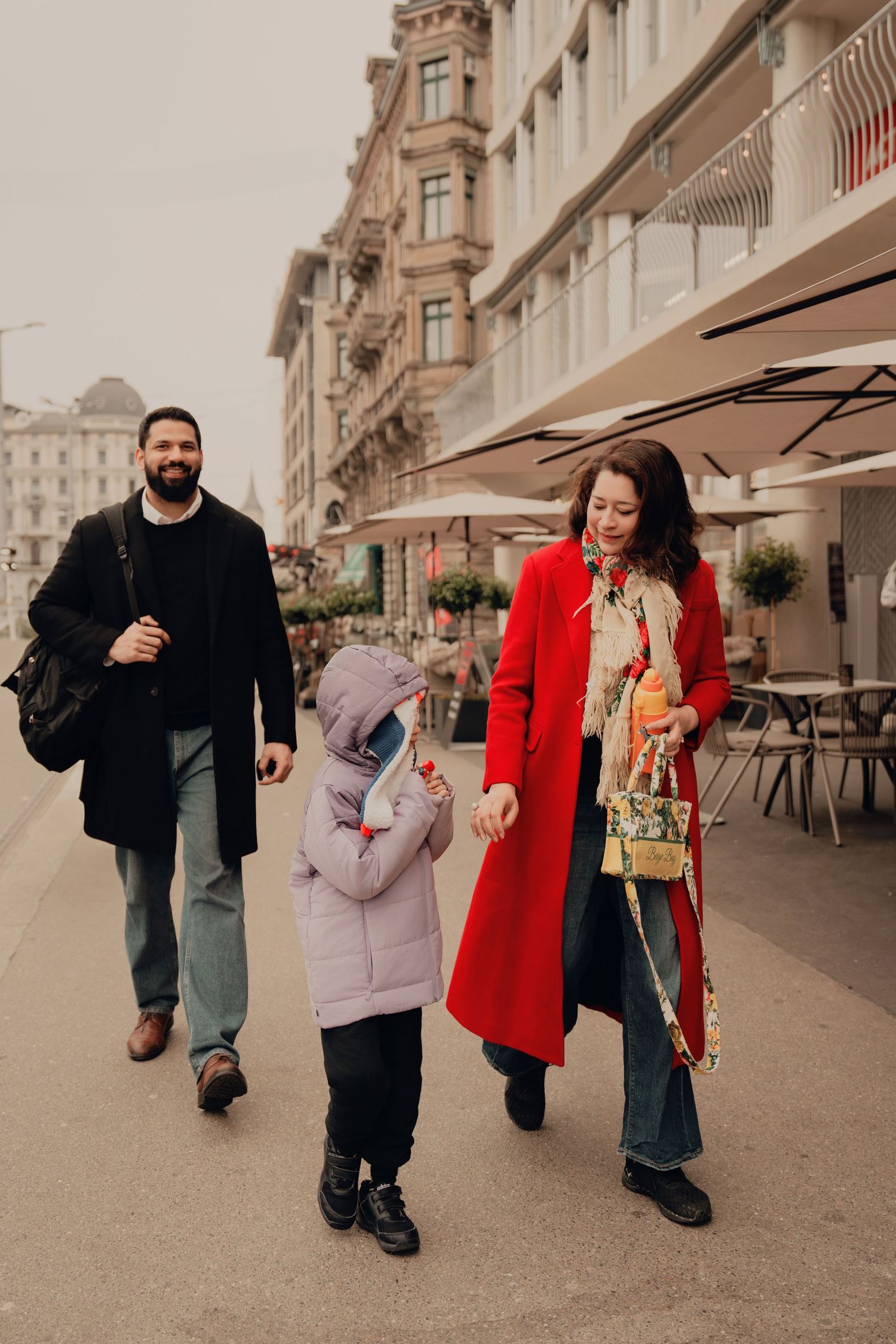 Smiling family of three walking together on European city sidewalk, child between parents in winter coats.