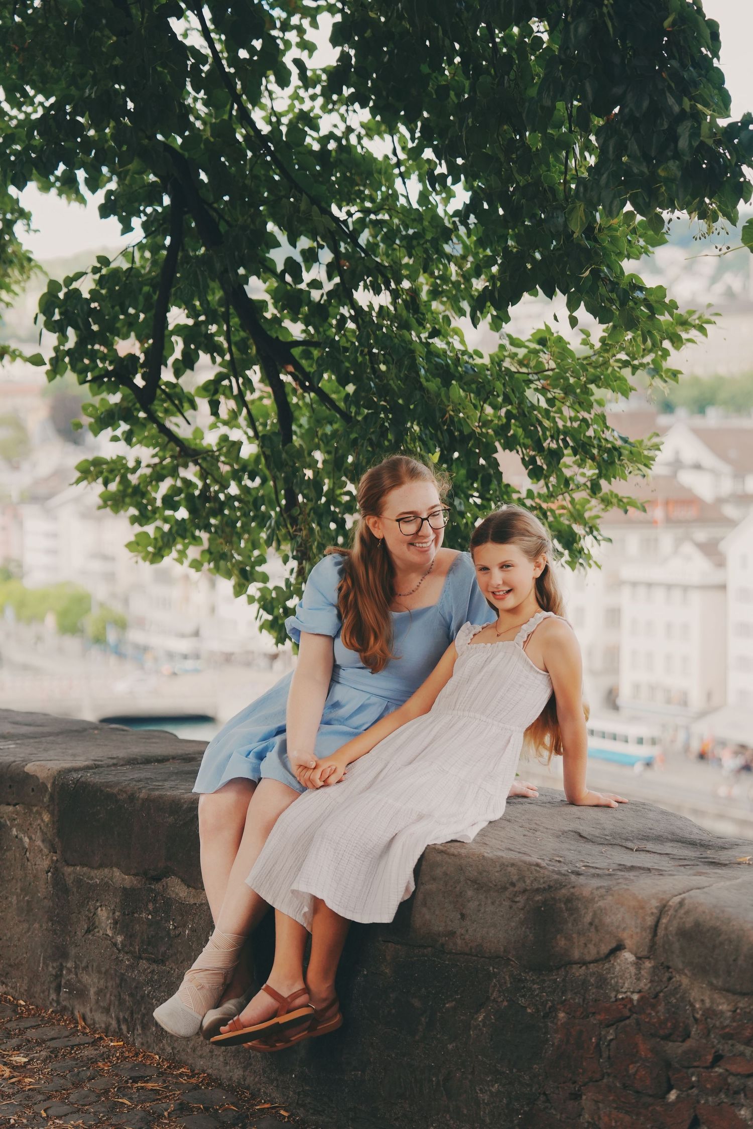 Mother and daughter sitting together on stone wall under large green tree in city.
