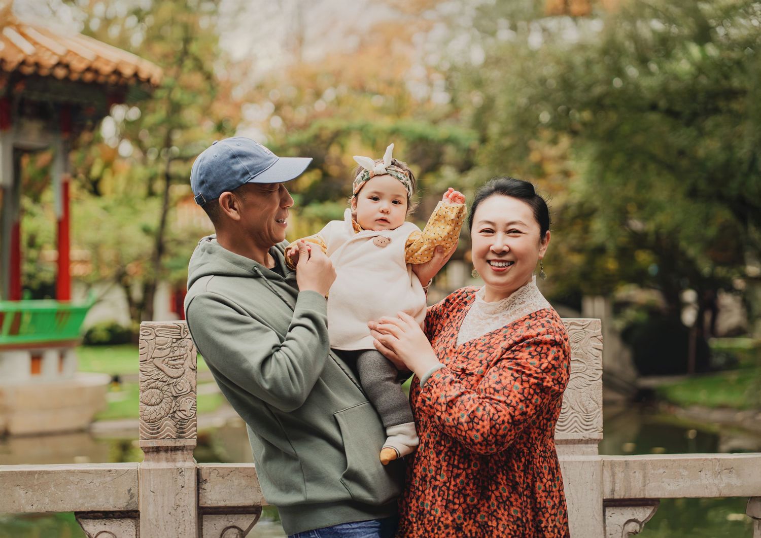 Grandparents smiling and holding baby together near stone railing in a Chinese garden.