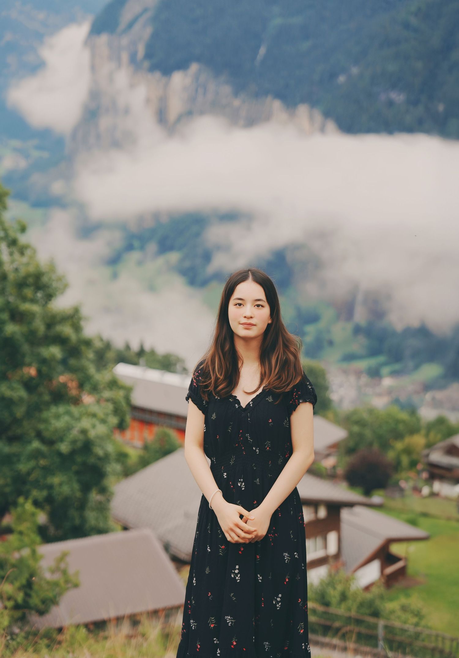 Young woman in black floral dress posing in front of scenic Swiss mountain valley with low clouds and chalets.
