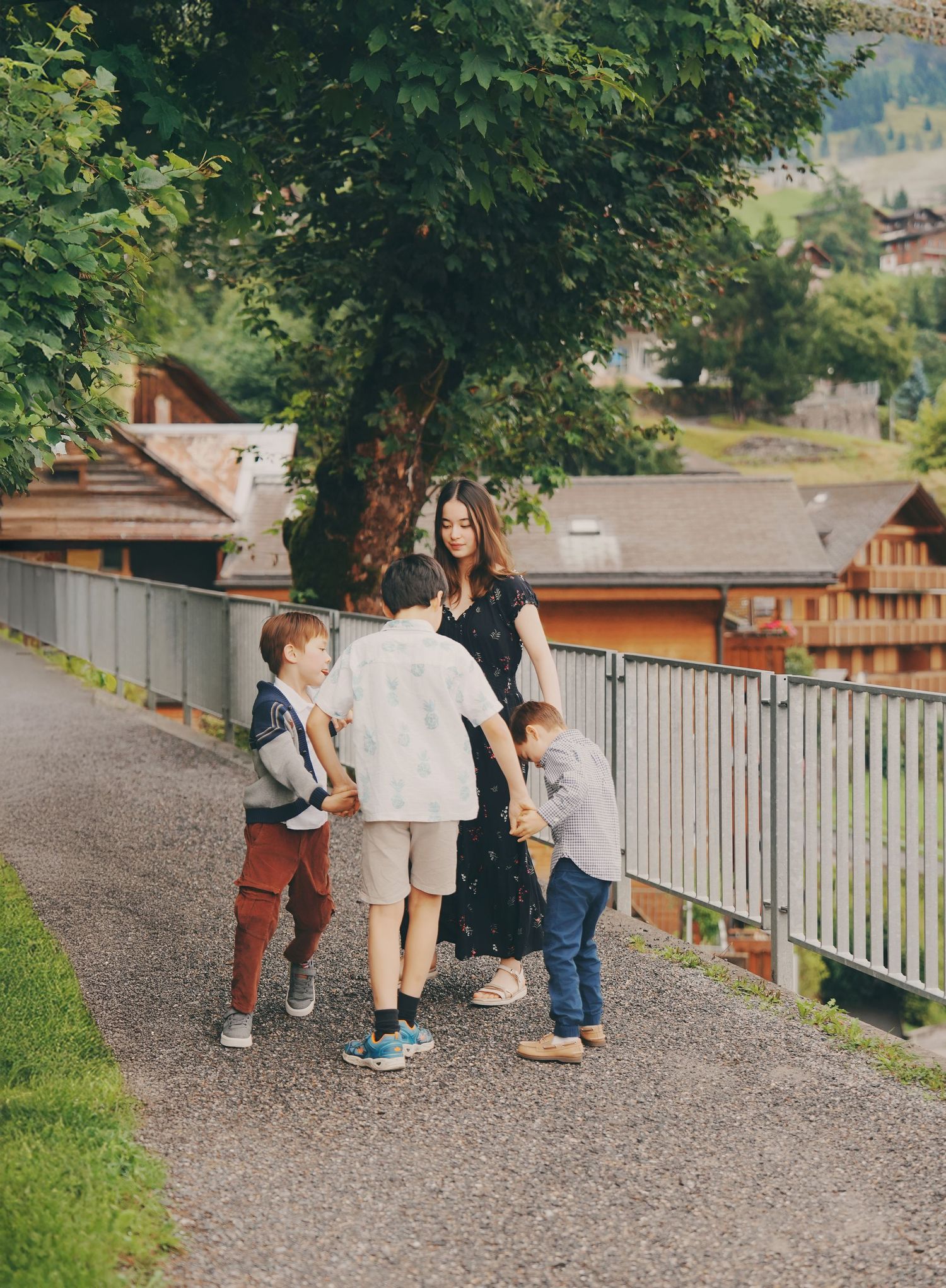 Woman in floral dress interacting playfully with three young boys on a tree-lined village pathway.