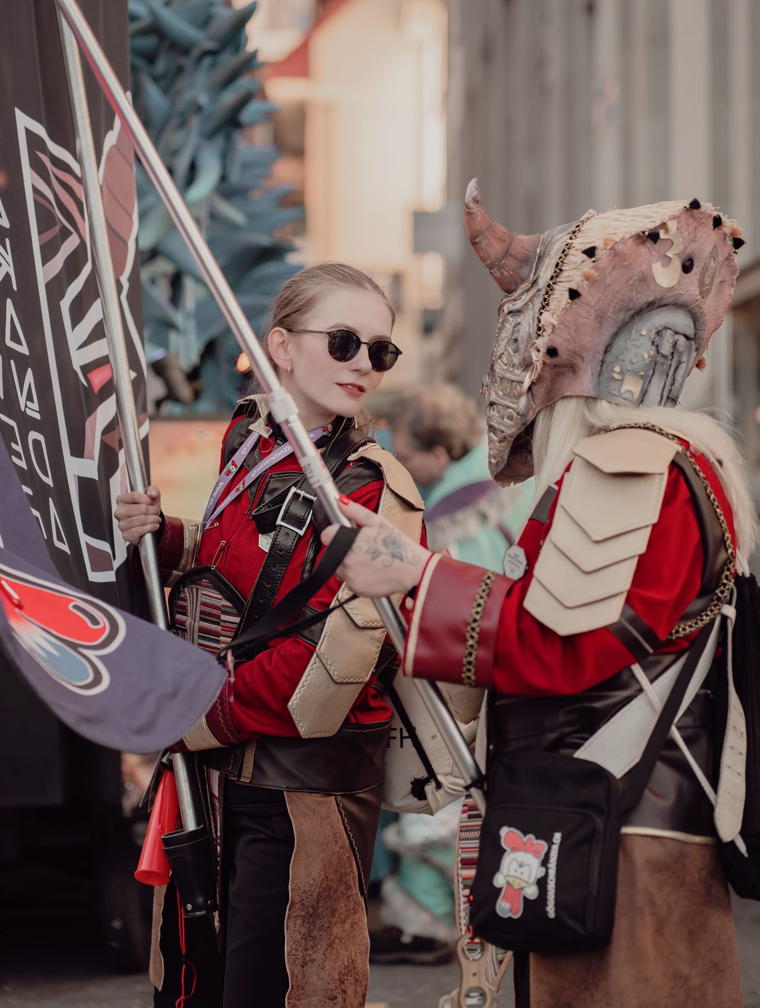 Two cosplayers in elaborate fantasy armor costumes holding flags at an outdoor convention event.