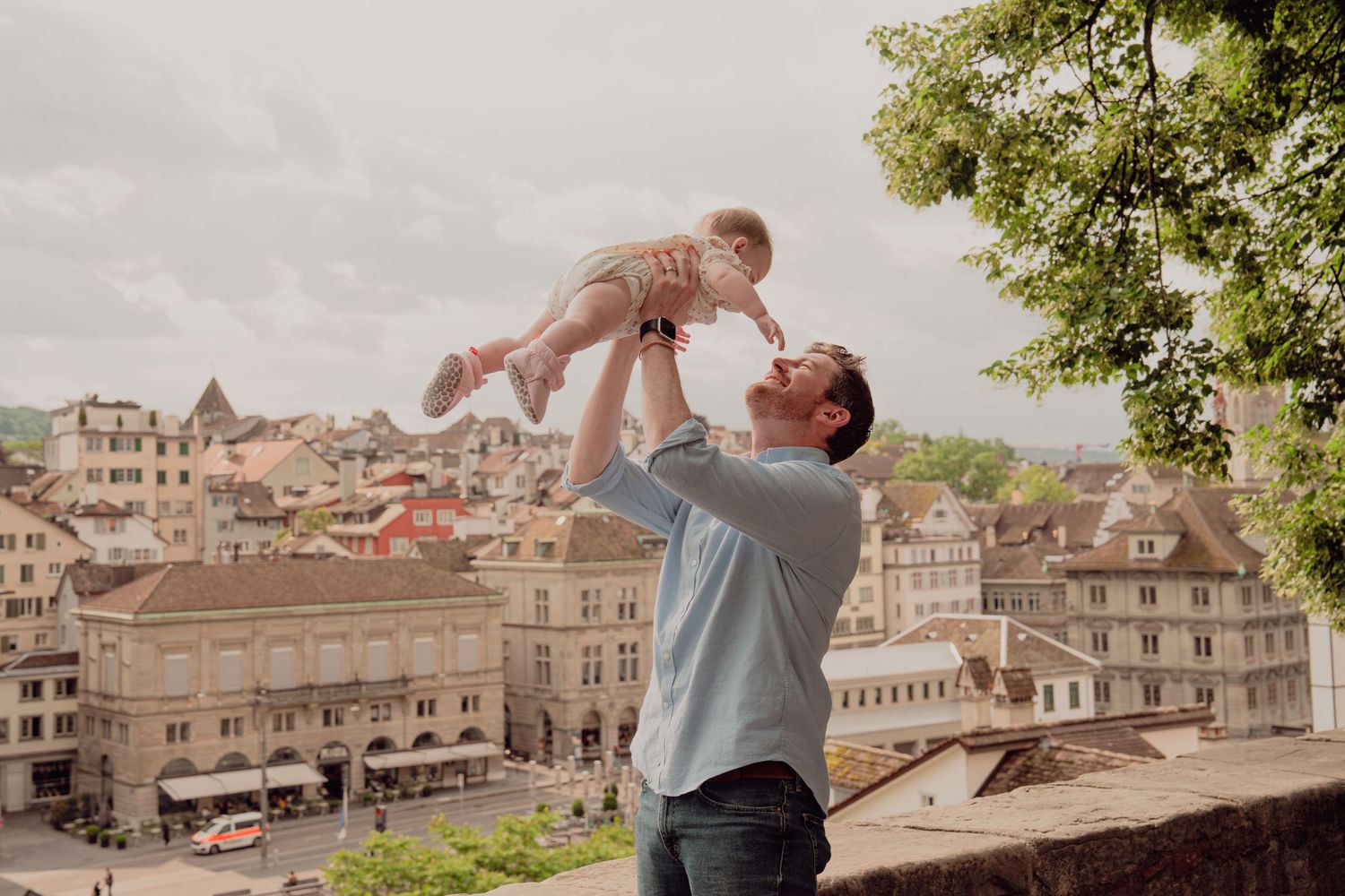 Father joyfully lifting baby in the air with European city skyline backdrop.