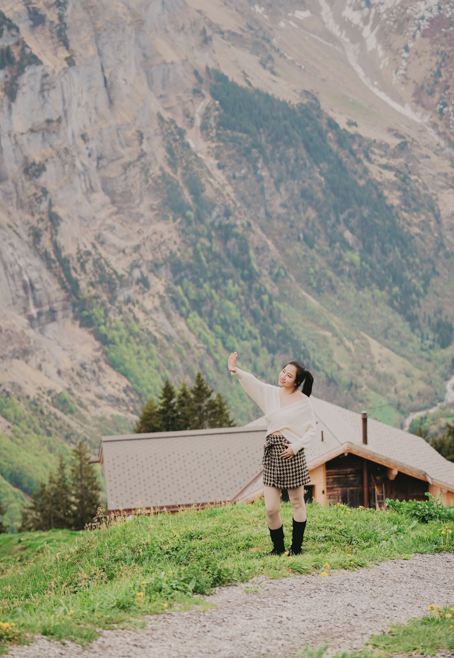 Young woman poses with outstretched arm on green alpine meadow with mountain backdrop and wooden cabin.