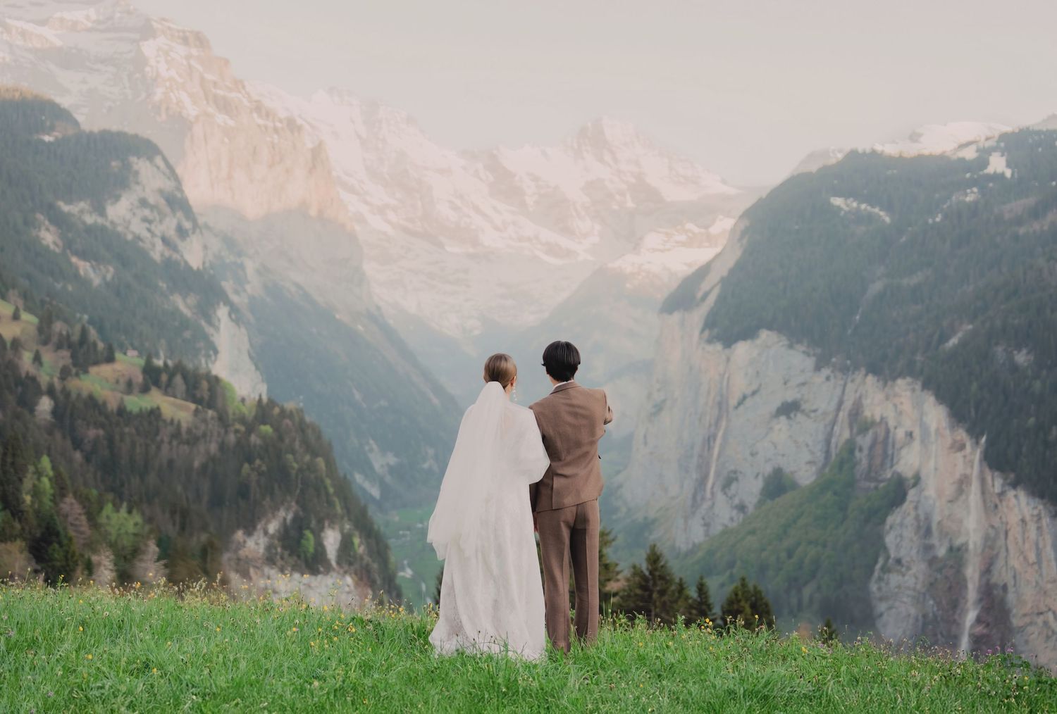 Wedding couple standing hand-in-hand on green meadow overlooking a dramatic misty Swiss mountain valley landscape.