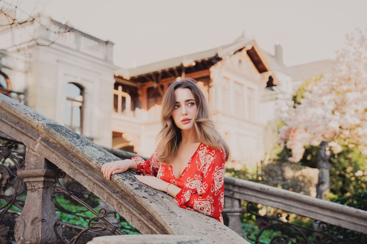 Young woman in red floral dress posing gracefully on stone steps near a historic building with blooming trees.