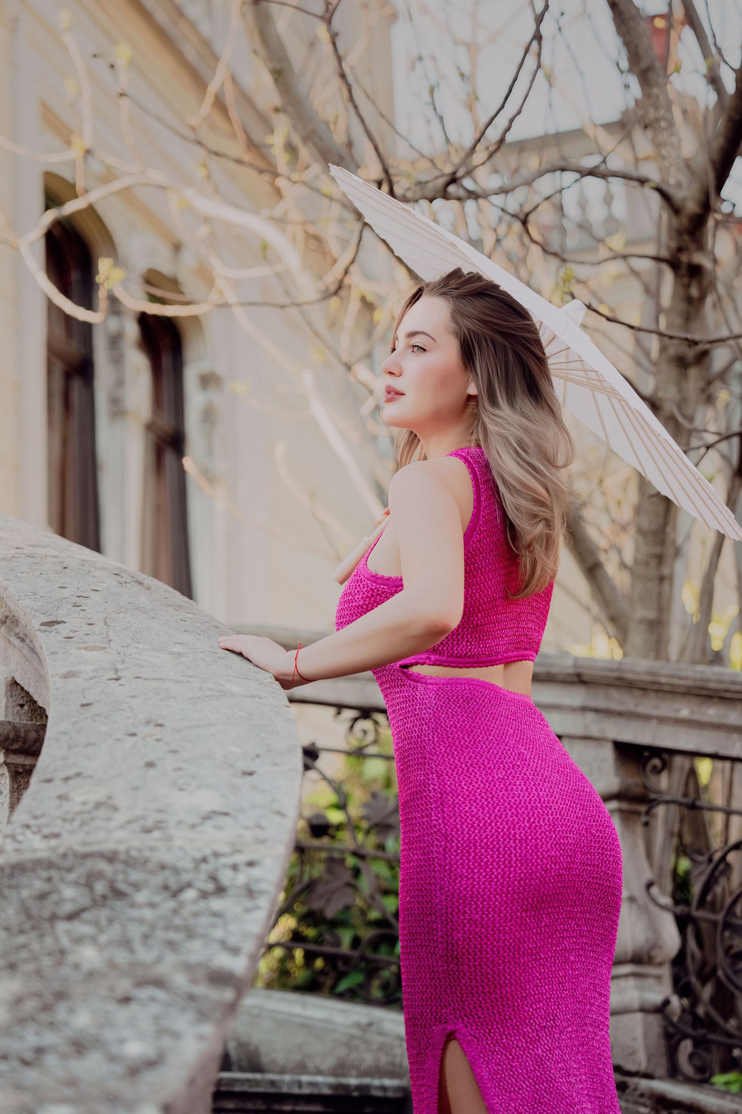 Woman in pink two-piece knit set leaning on stone balustrade holding parasol near historic building.