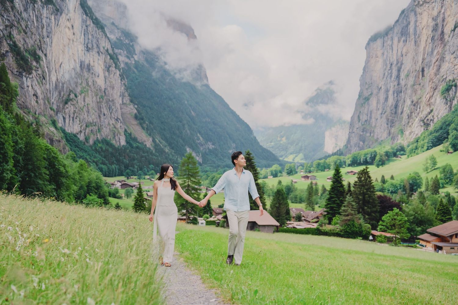 Couple walking hand in hand through a lush green Swiss alpine meadow with dramatic mountain scenery.