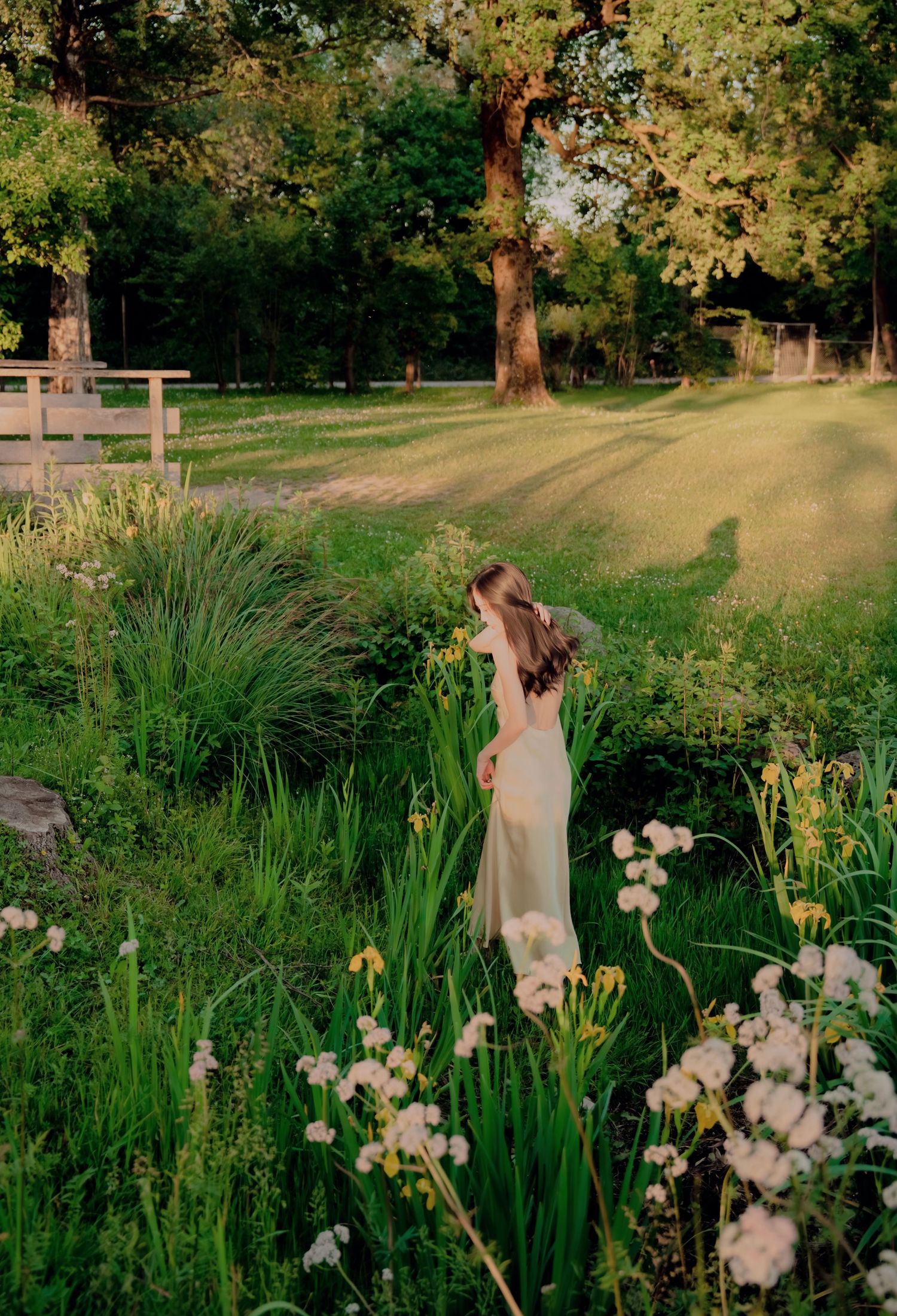 Woman in pale yellow dress standing among wildflowers and lush greenery in a sunlit garden at golden hour.