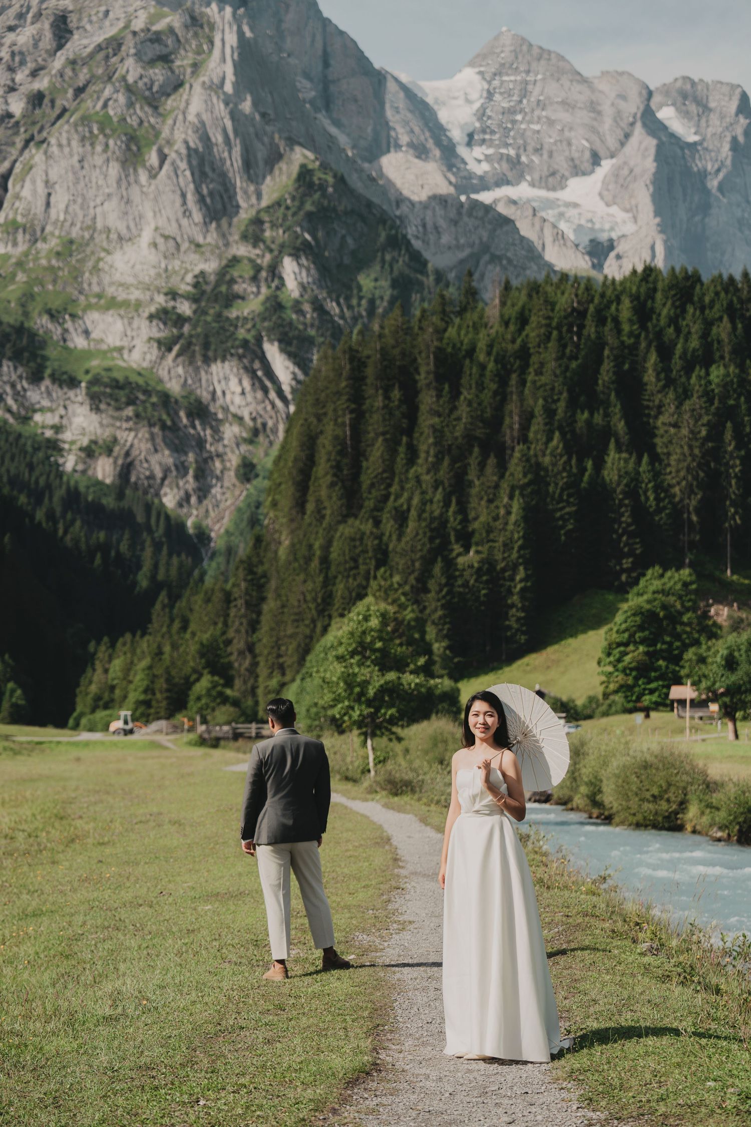 Bride in white gown holding parasol stands on alpine path with groom, majestic Swiss mountain backdrop.