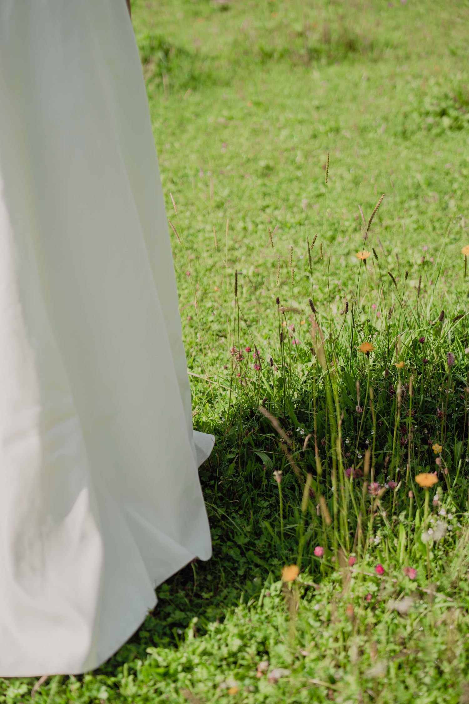 Detail shot of white bridal gown train on vibrant green grass with colorful wildflowers.