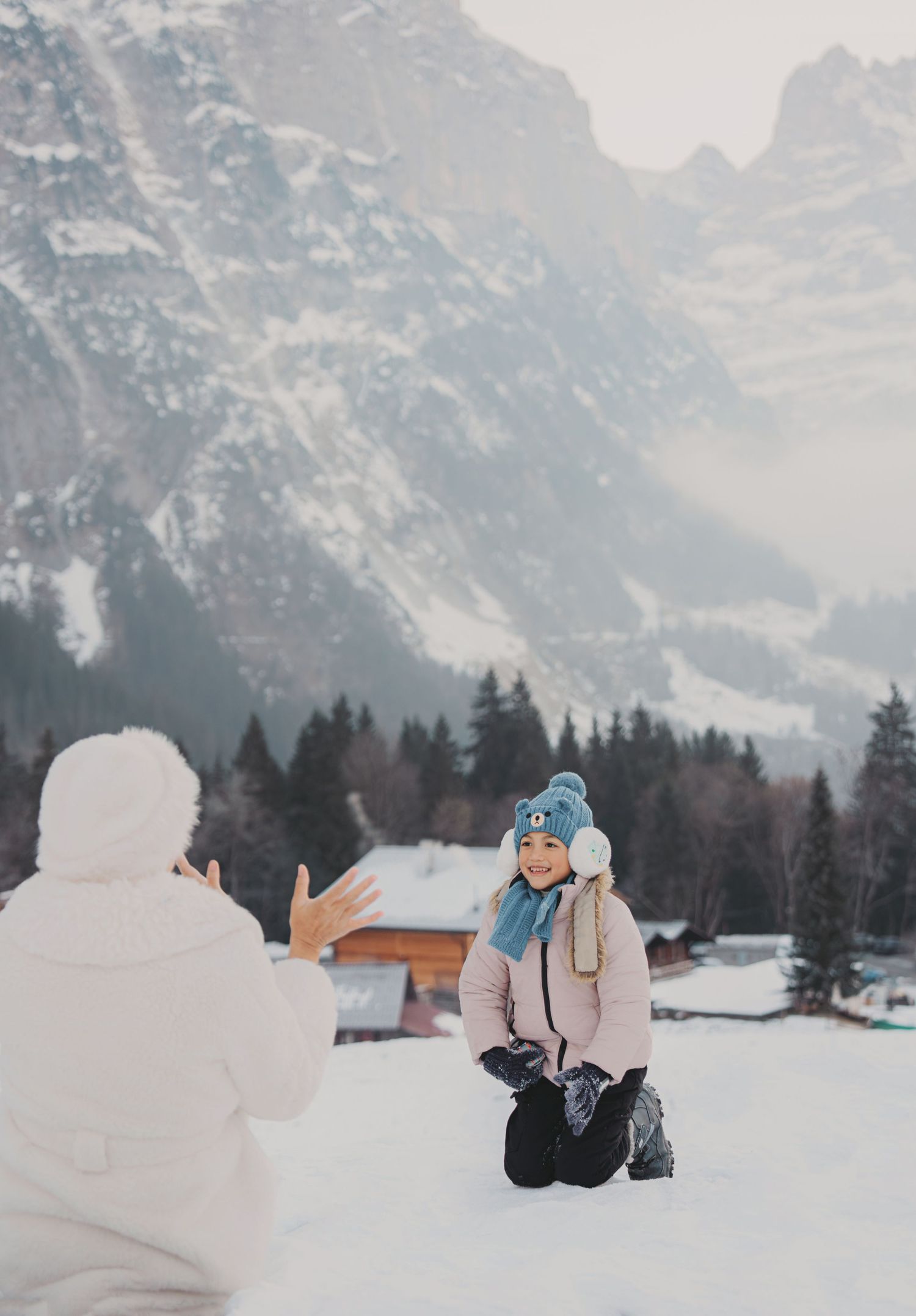 Young girl in blue knit hat smiling while playing in snow with alpine mountains and forest behind her.