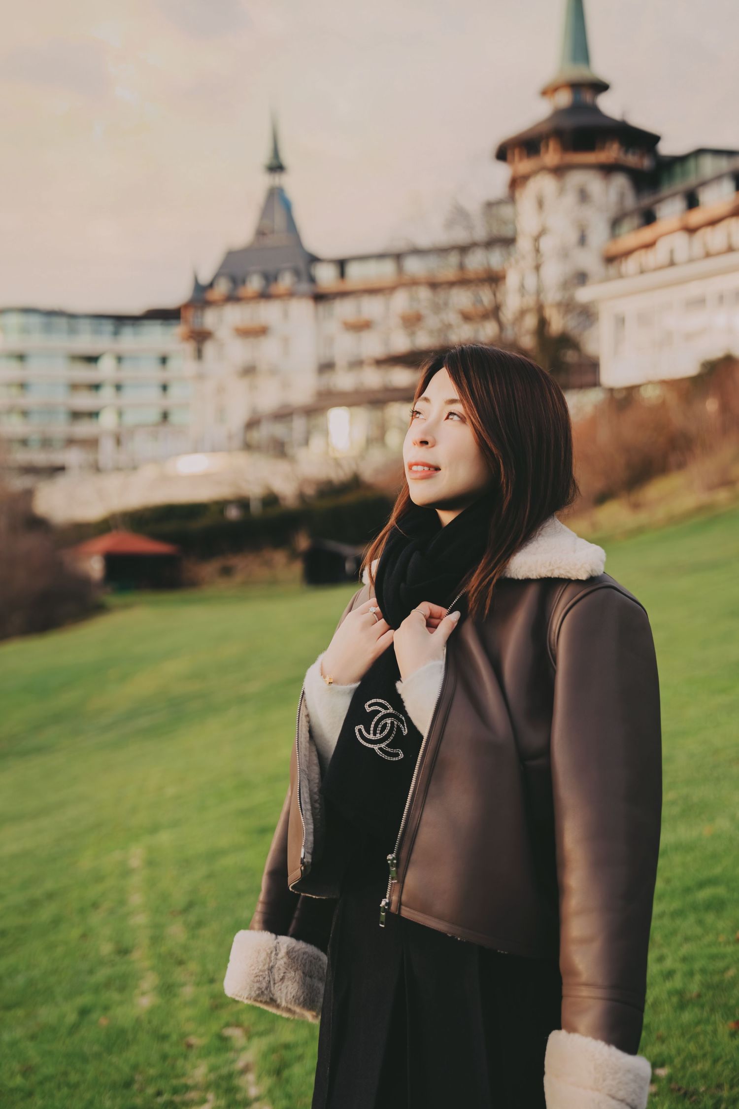 Stylish Asian woman in Chanel shearling coat posing before a European castle on green lawn.