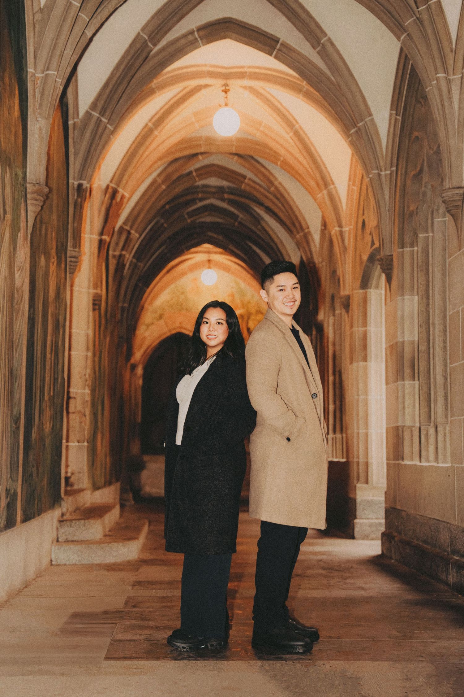 Engaged couple standing back-to-back smiling in a stunning Gothic arched stone hallway.