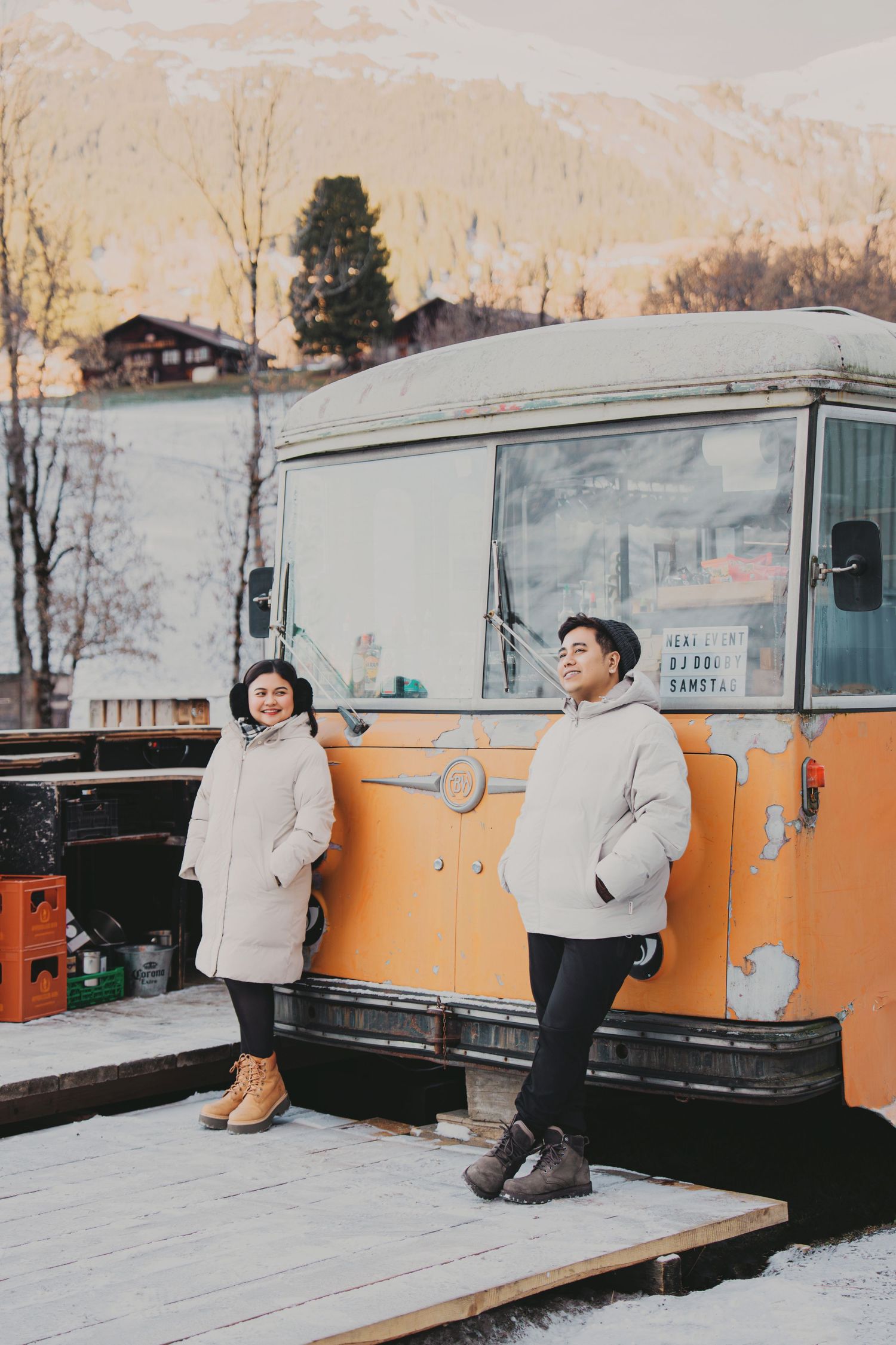 Couple in white winter coats posing beside vintage orange food truck on snowy mountain platform.