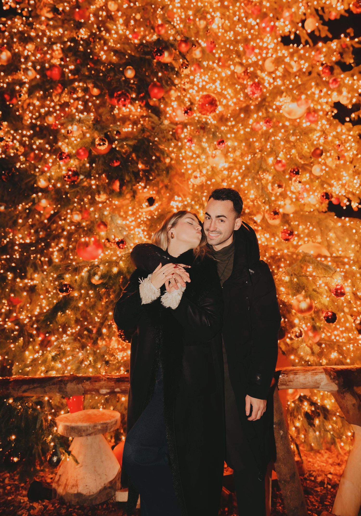 Couple posing affectionately in front of a stunning illuminated Christmas tree decorated with red ornaments.