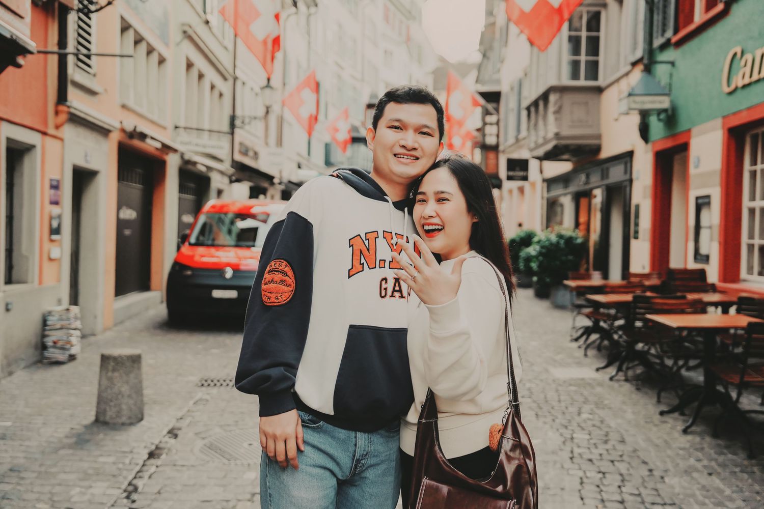 Smiling Asian couple posing together on colorful European cobblestone street with Swiss flags.